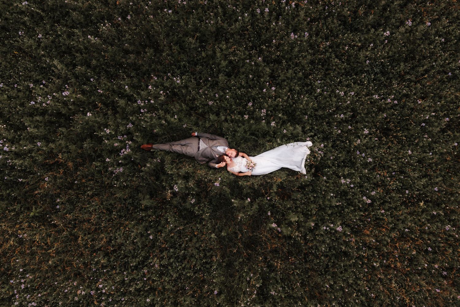 Aerial drone view of bride and groom lying in a green field with white wedding dress flowing.