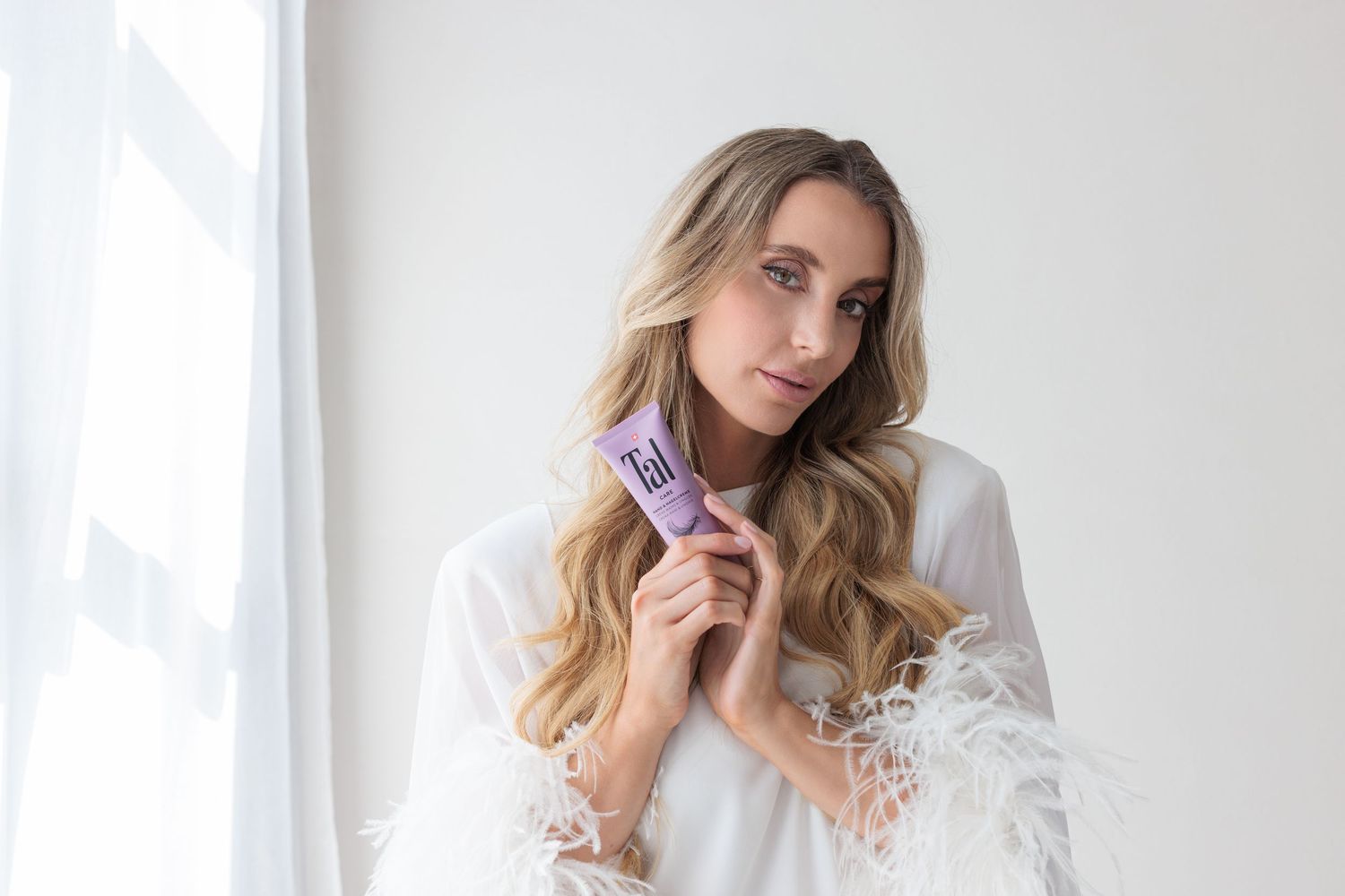 Blonde woman in white feathered robe holding purple Tal skincare tube near bright window.