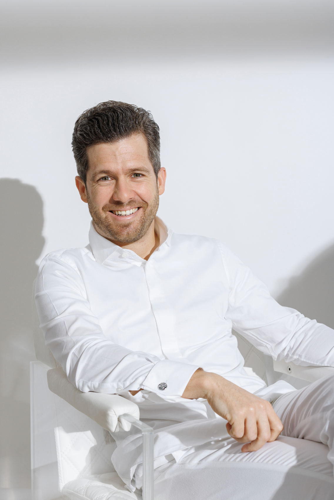 A smiling man in a white dress shirt sits relaxed in a modern chair against a bright white background.