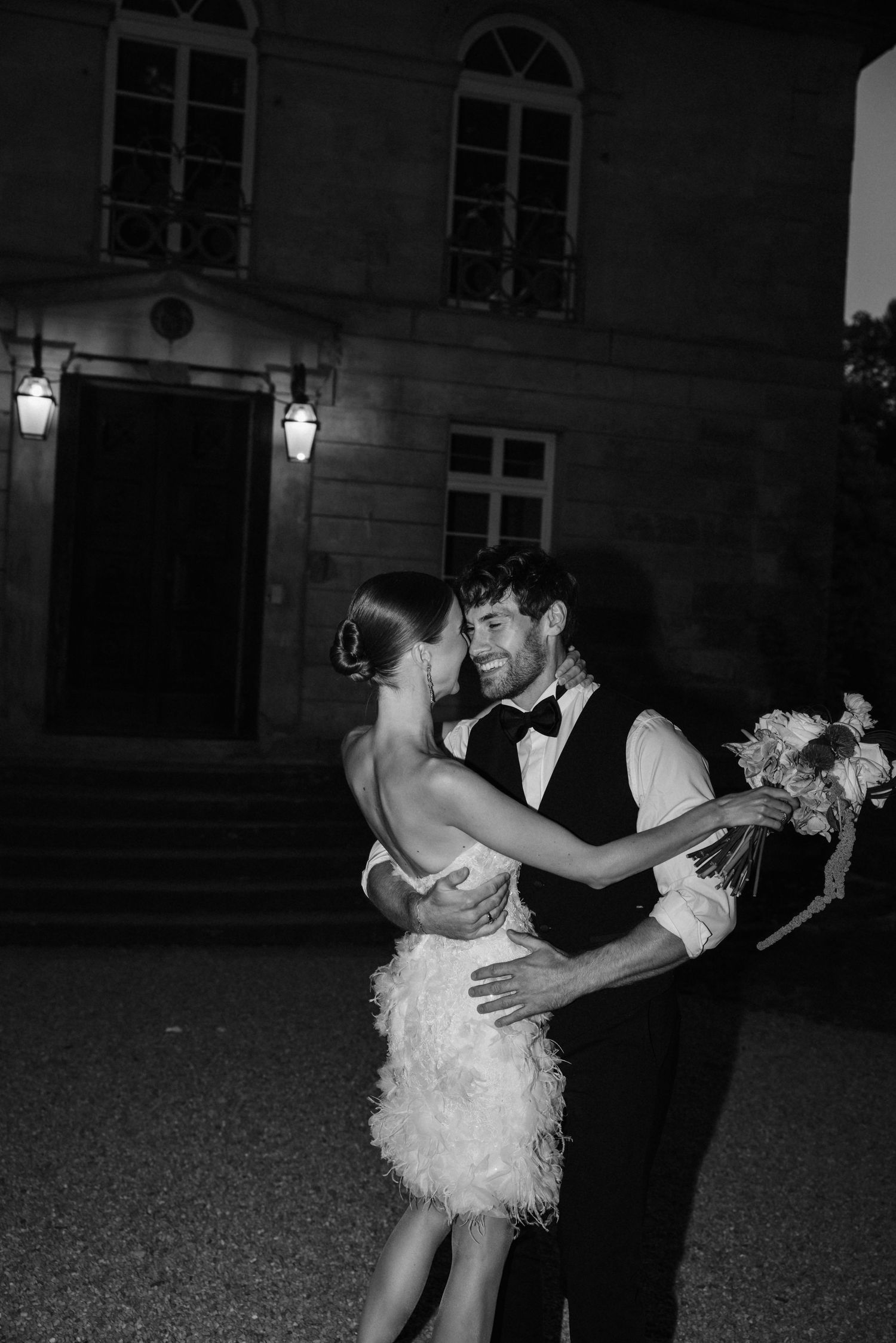 Black and white nighttime photo of bride in feather mini dress and groom dancing outside estate.