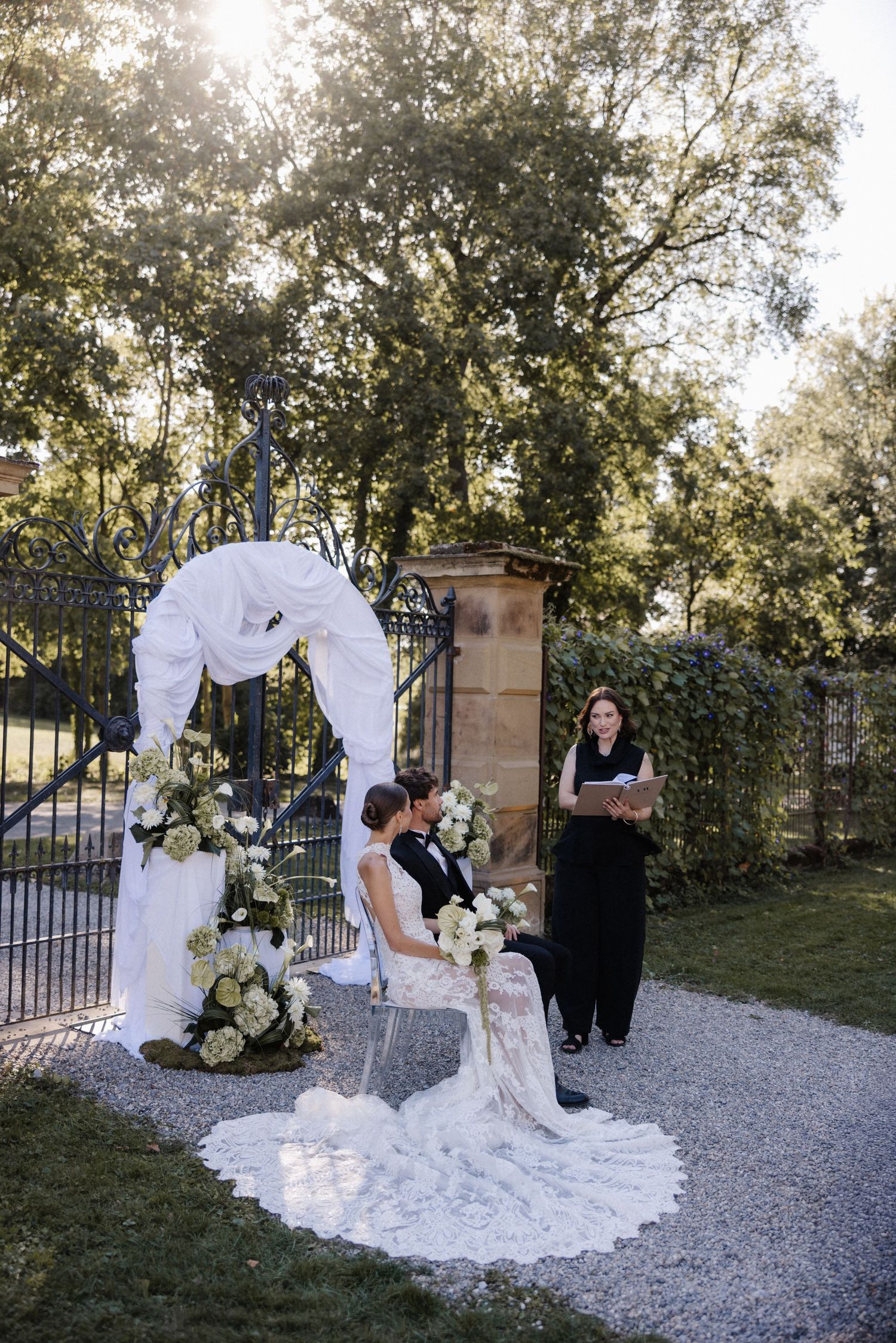 Bride and groom sit beneath a white draped floral arch during an outdoor wedding ceremony.