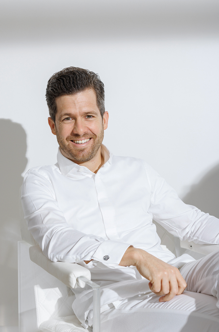 Smiling man in white dress shirt sitting relaxed on white chair against light background.