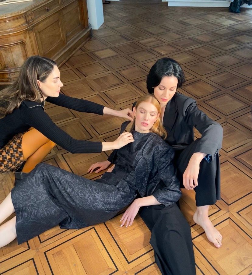 Three women in black outfits posed dramatically on a parquet floor in an elegant interior.