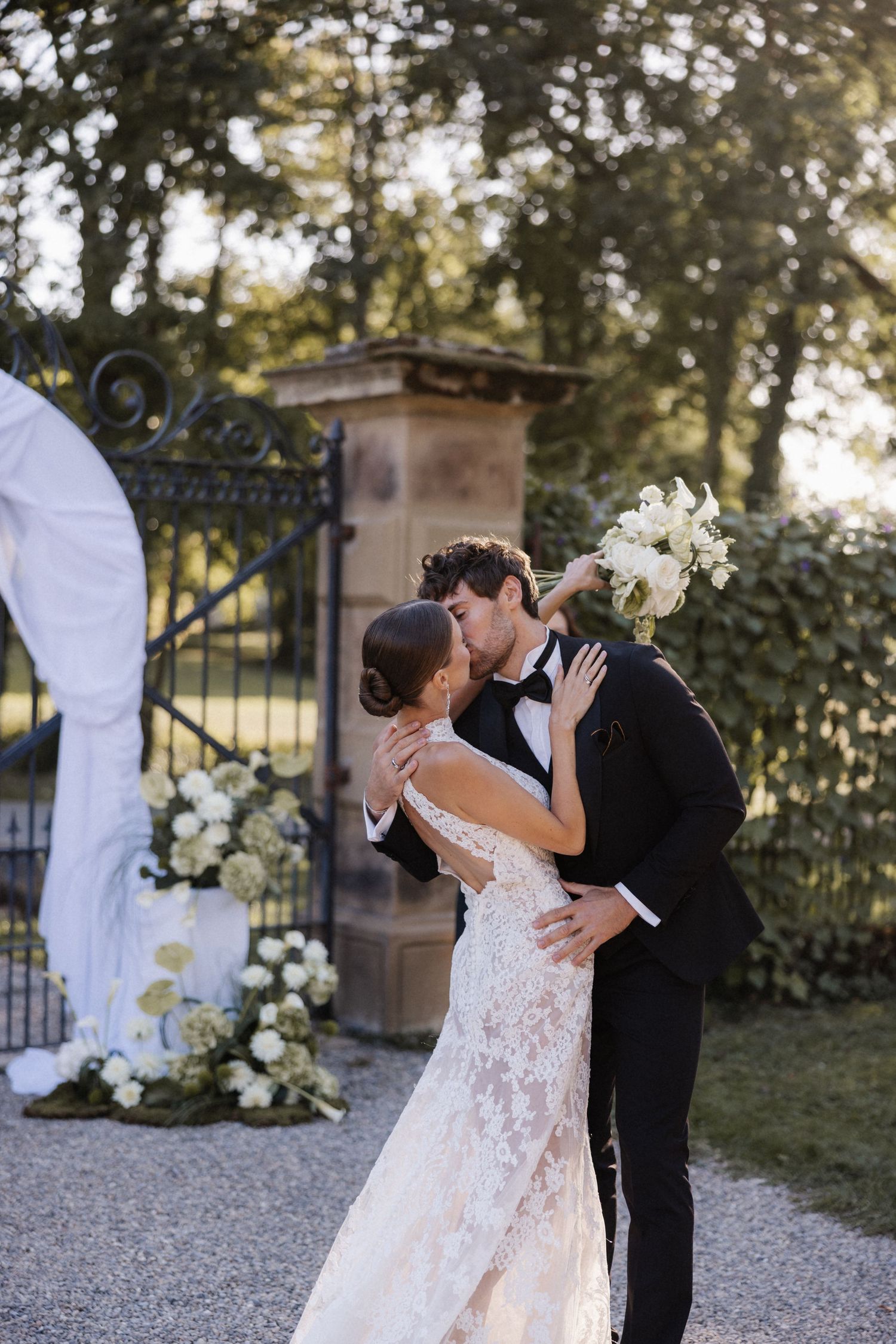Bride and groom share a romantic kiss outdoors near a white draped arch with white floral arrangements.