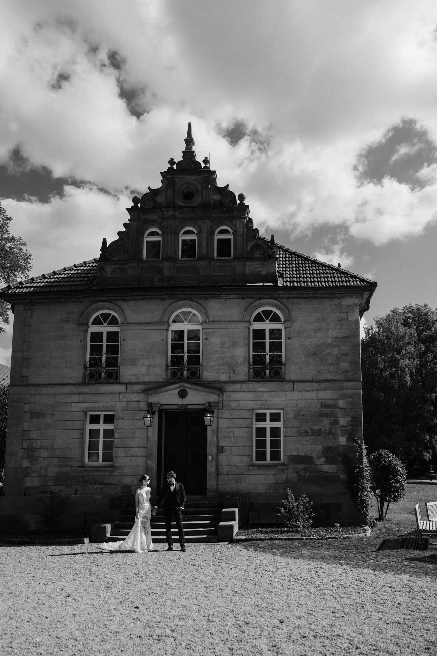 Black and white photo of bride and groom standing before an ornate historic stone manor house.
