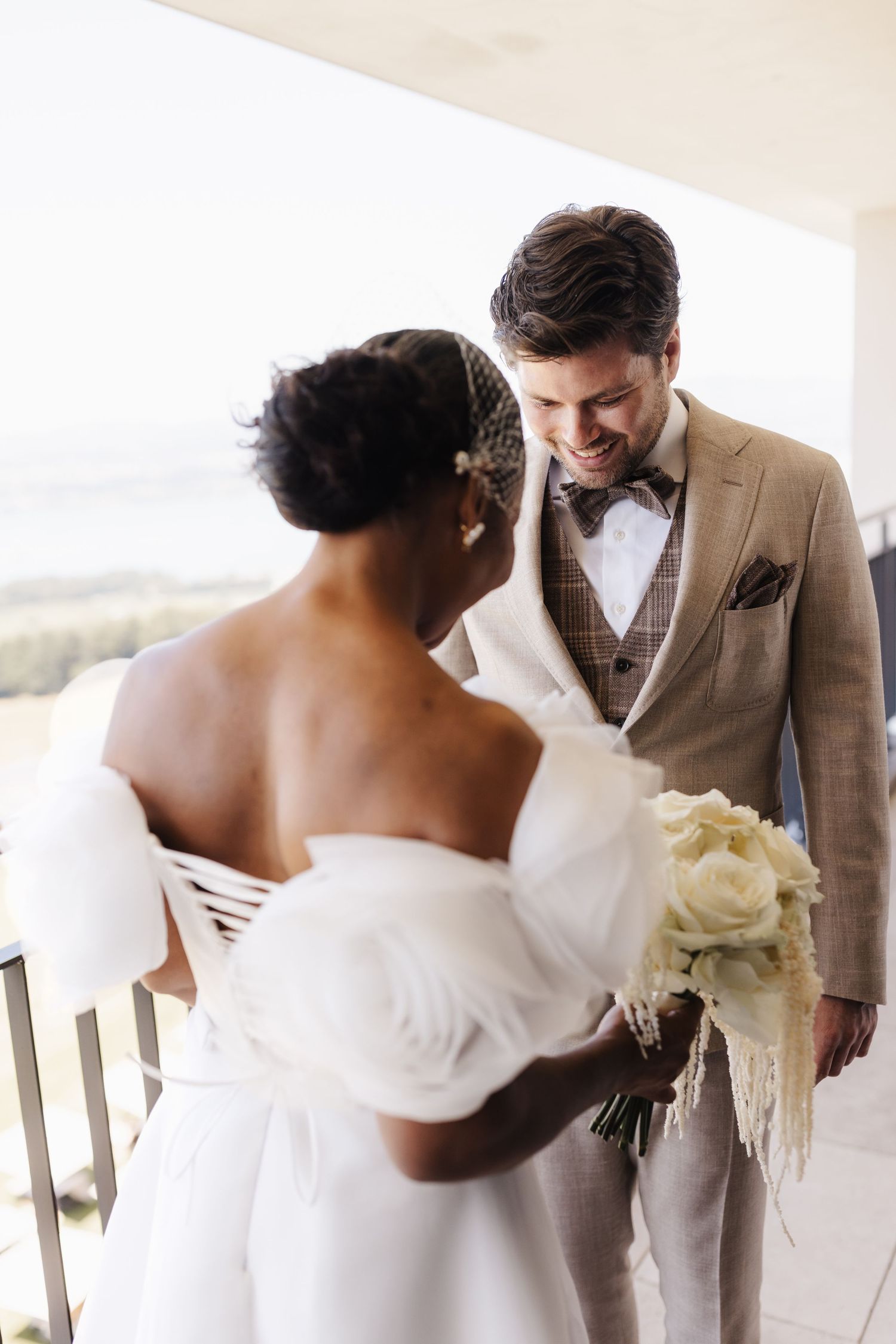 Groom in tan suit smiles at Black bride in white off-shoulder gown during outdoor first look.