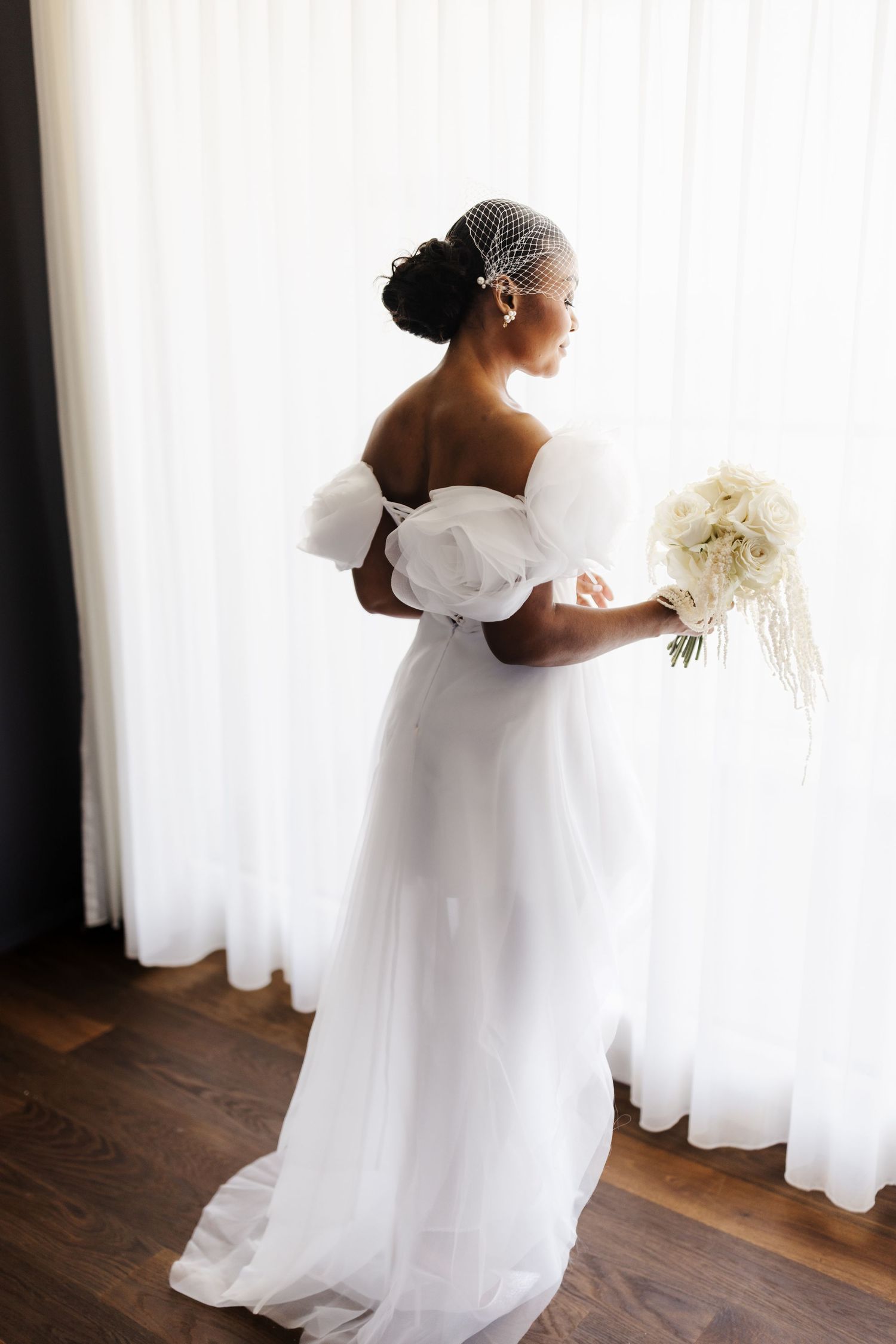 Black bride in white off-shoulder tulle gown and birdcage veil holds white bouquet by window.