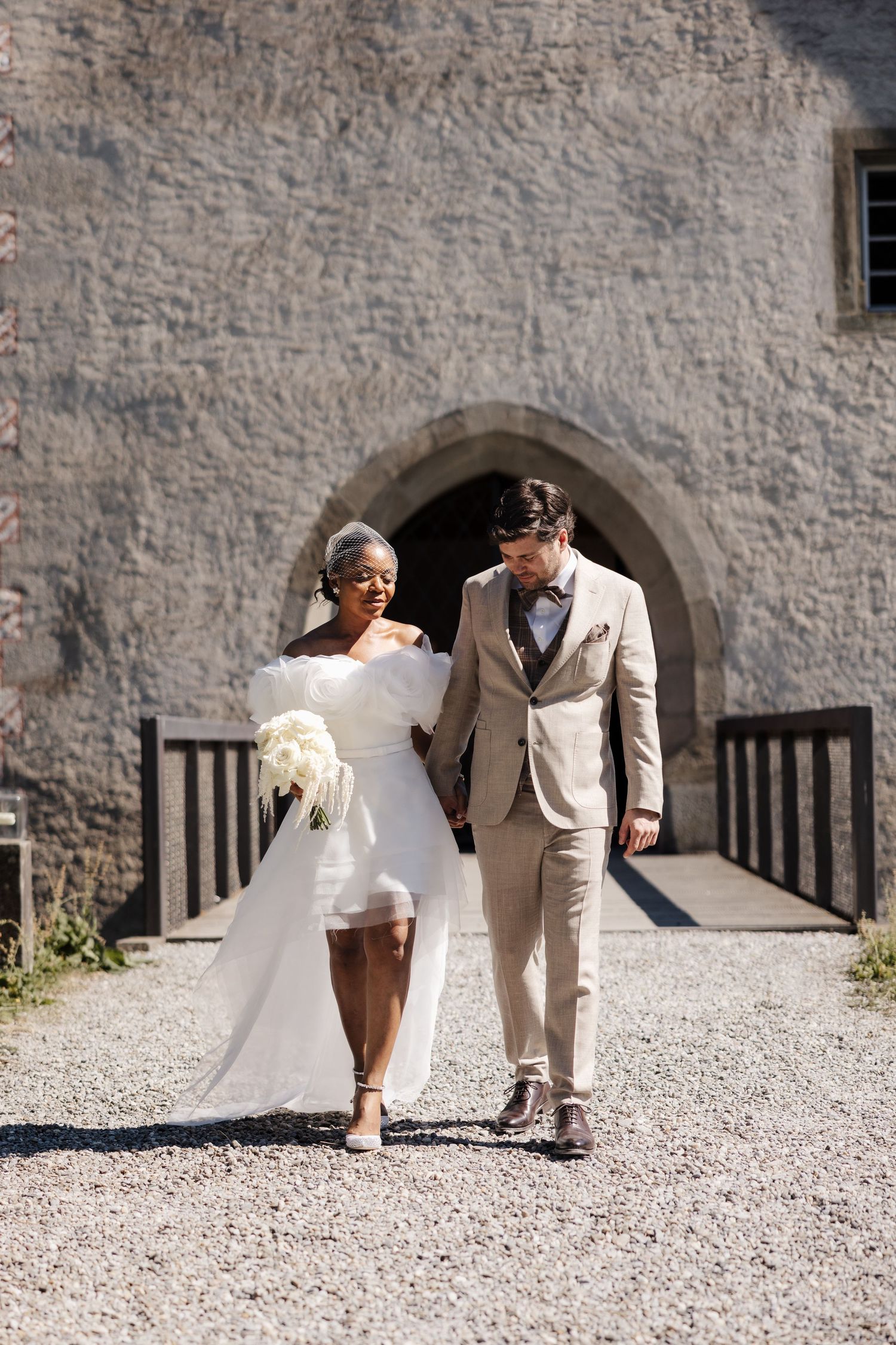 Smiling interracial bride and groom walking hand in hand outside a rustic stone castle venue.