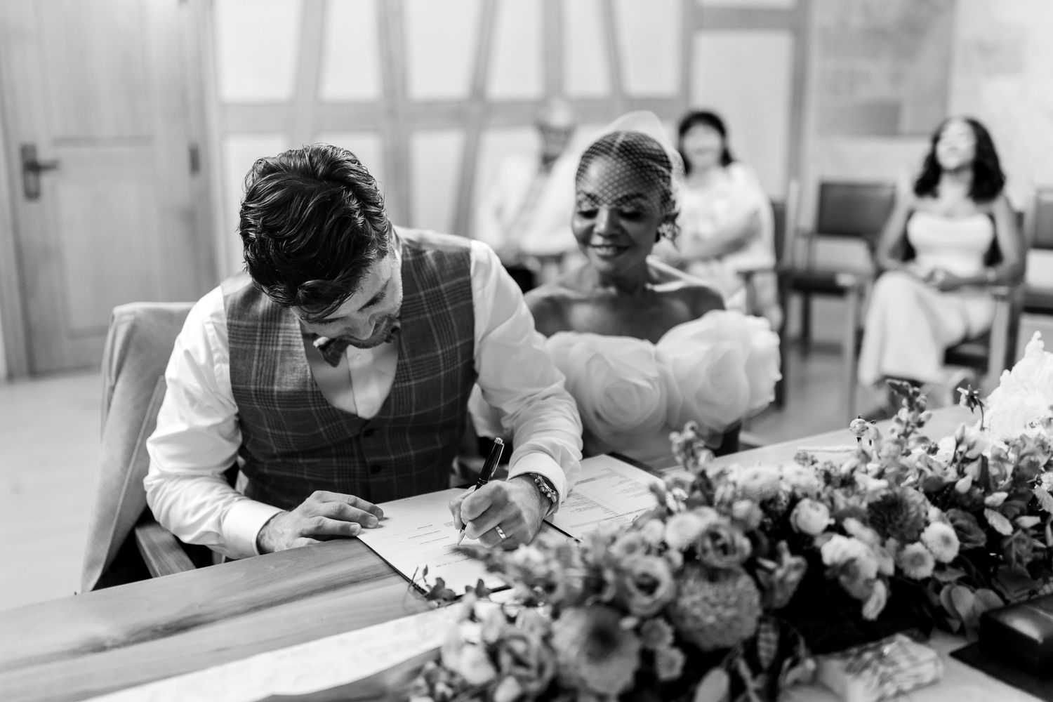 Black and white photo of groom signing wedding register as smiling bride watches at ceremony.