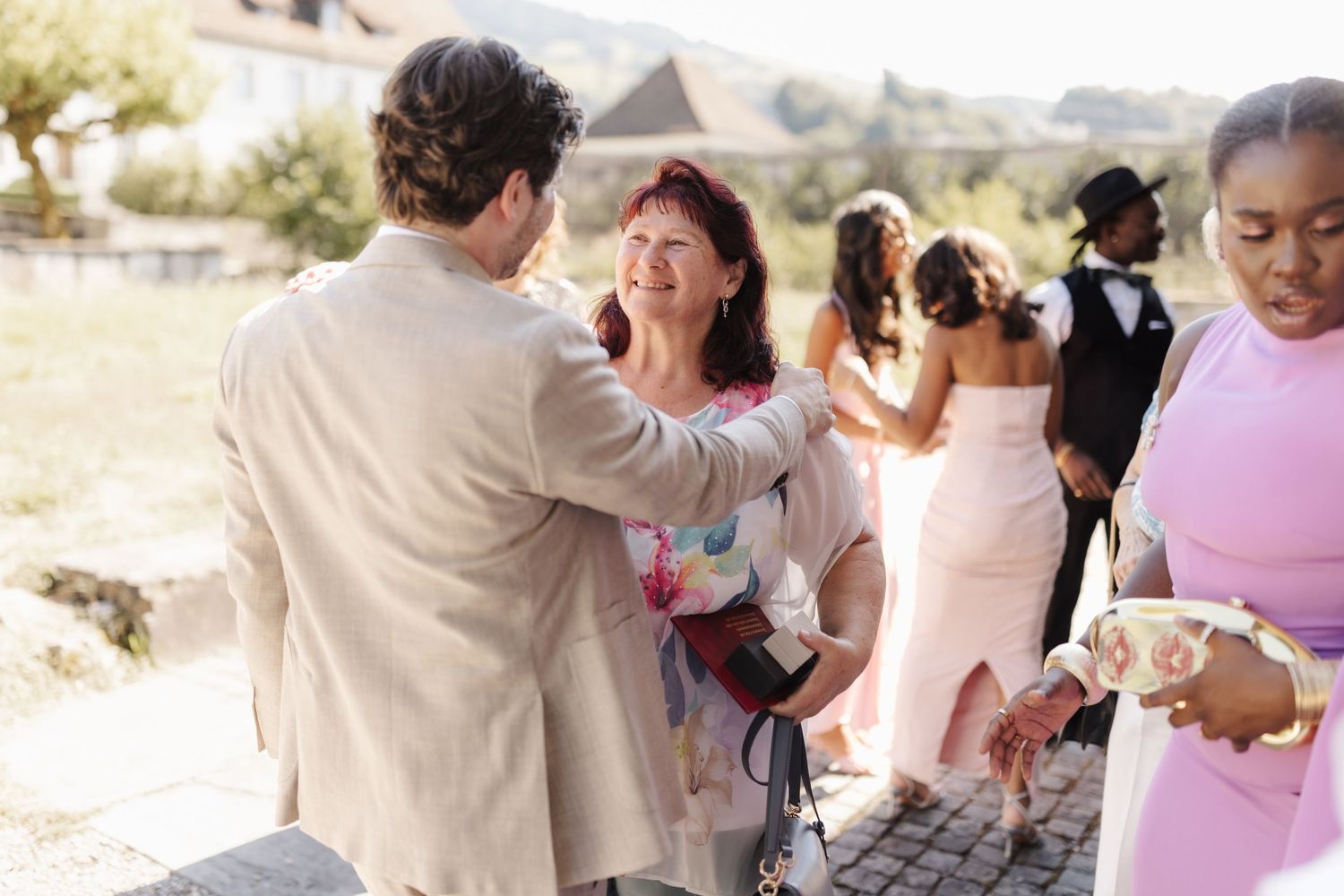 Groom in beige suit greets smiling red-haired woman at outdoor wedding reception with guests mingling behind.