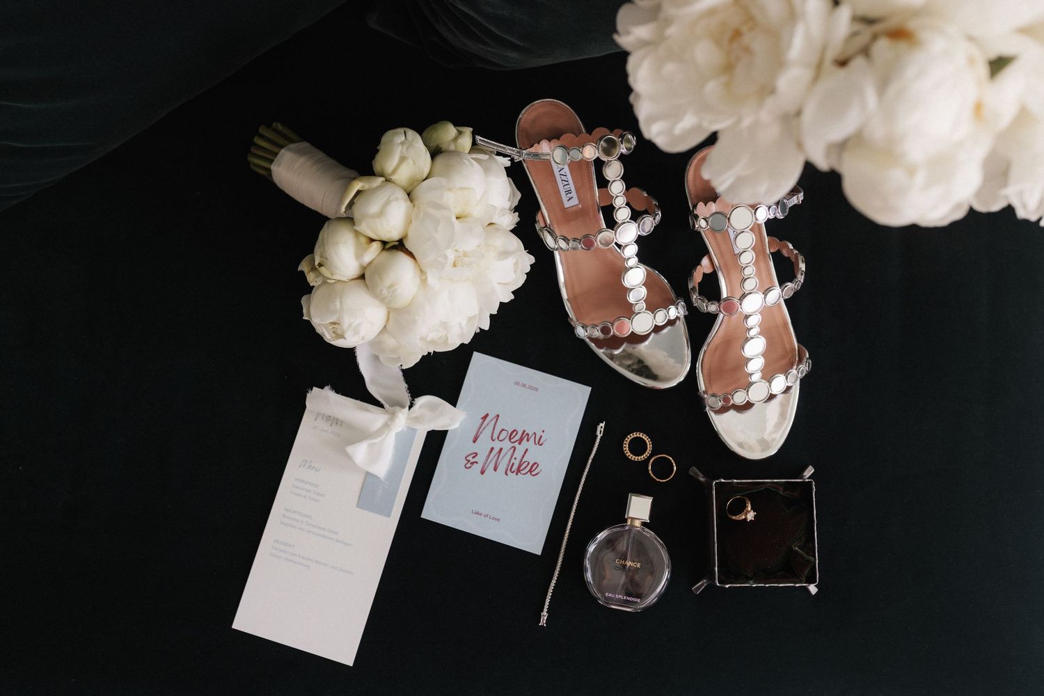Flat lay of bridal accessories including shoes, peonies, rings, and wedding invitation.