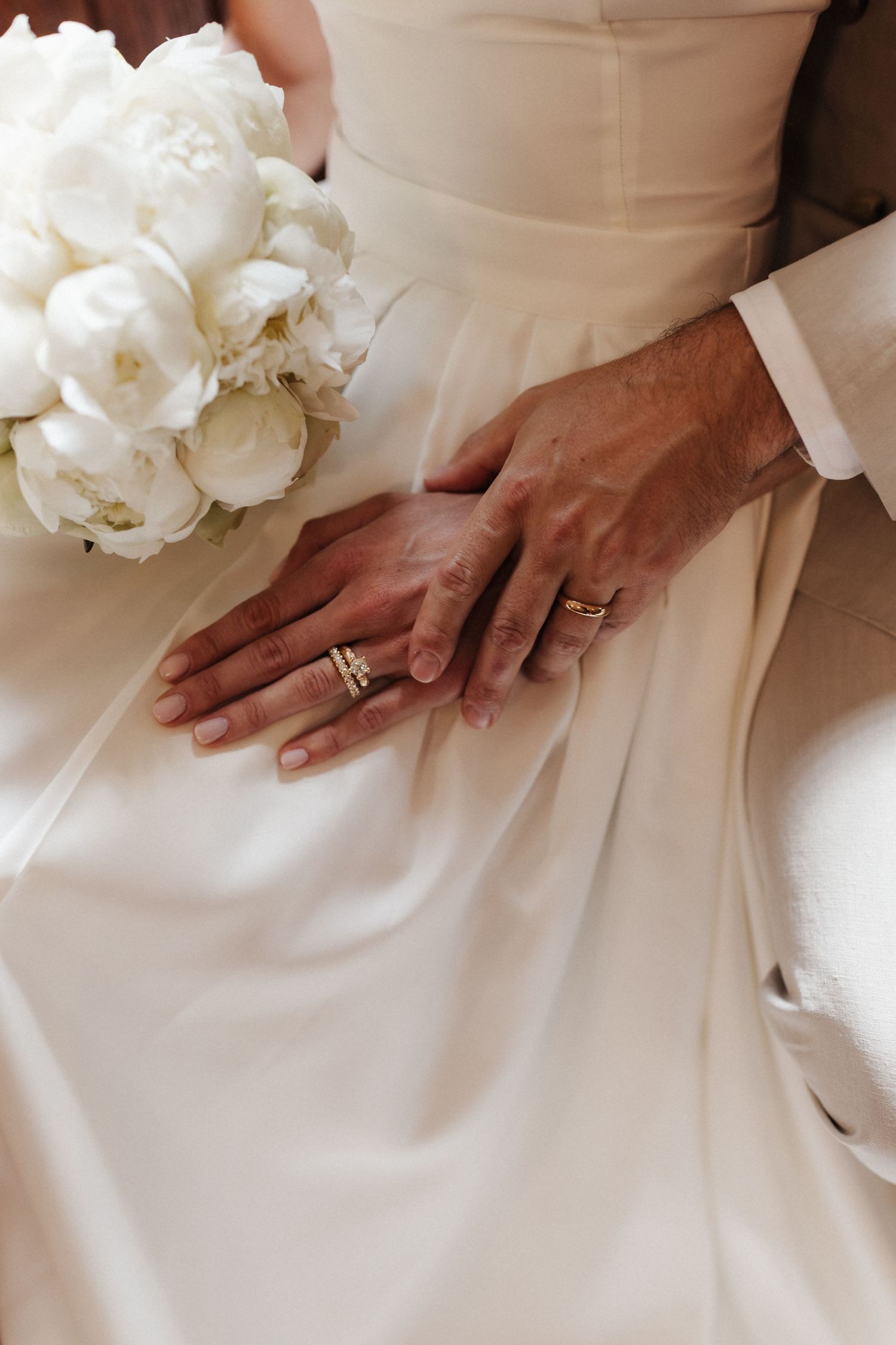 Close-up of newlyweds' hands showing gold wedding rings resting on bride's white gown beside white peony bouquet.