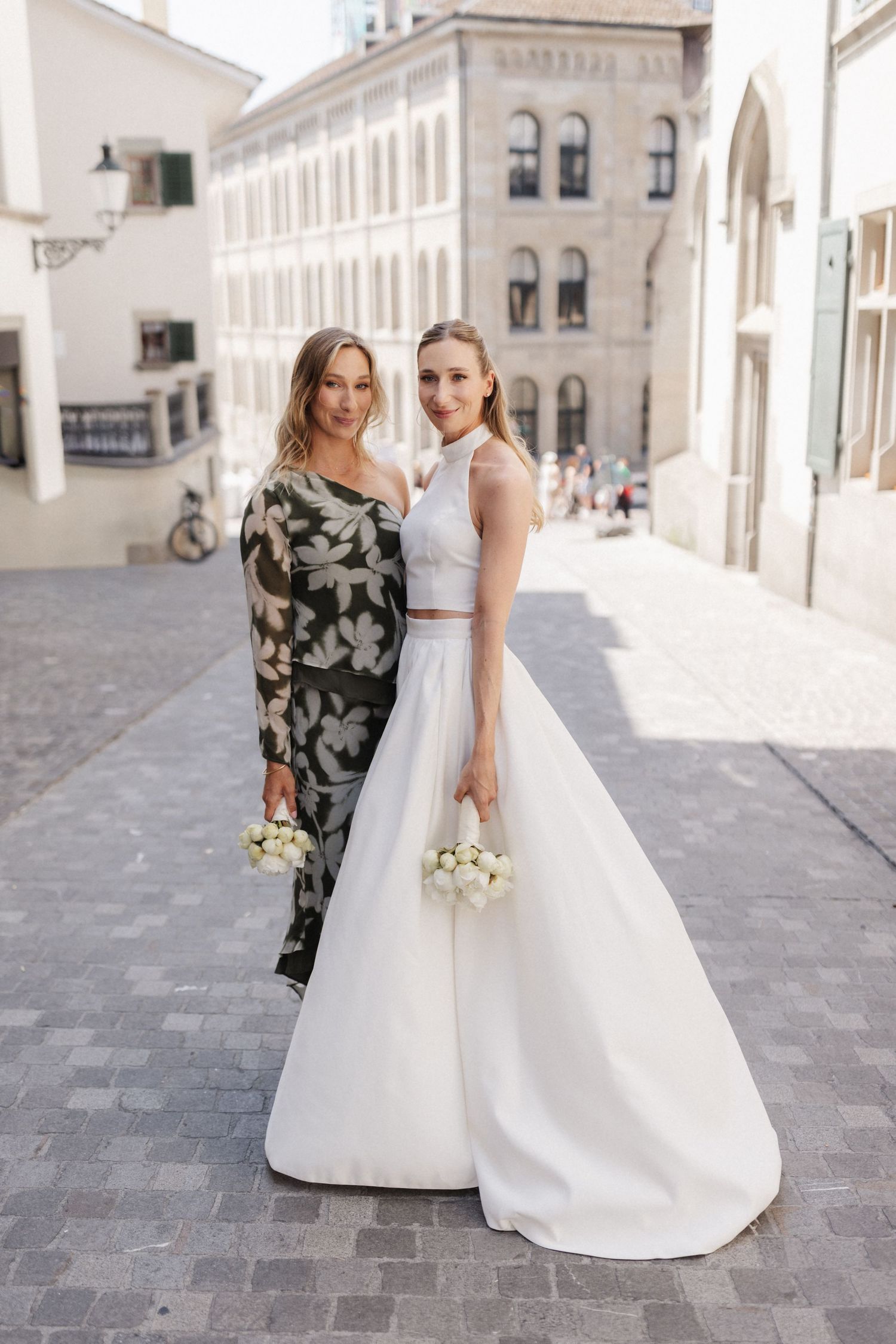 A bride in a white halter gown and a woman in a floral dress pose together on a cobblestone street.