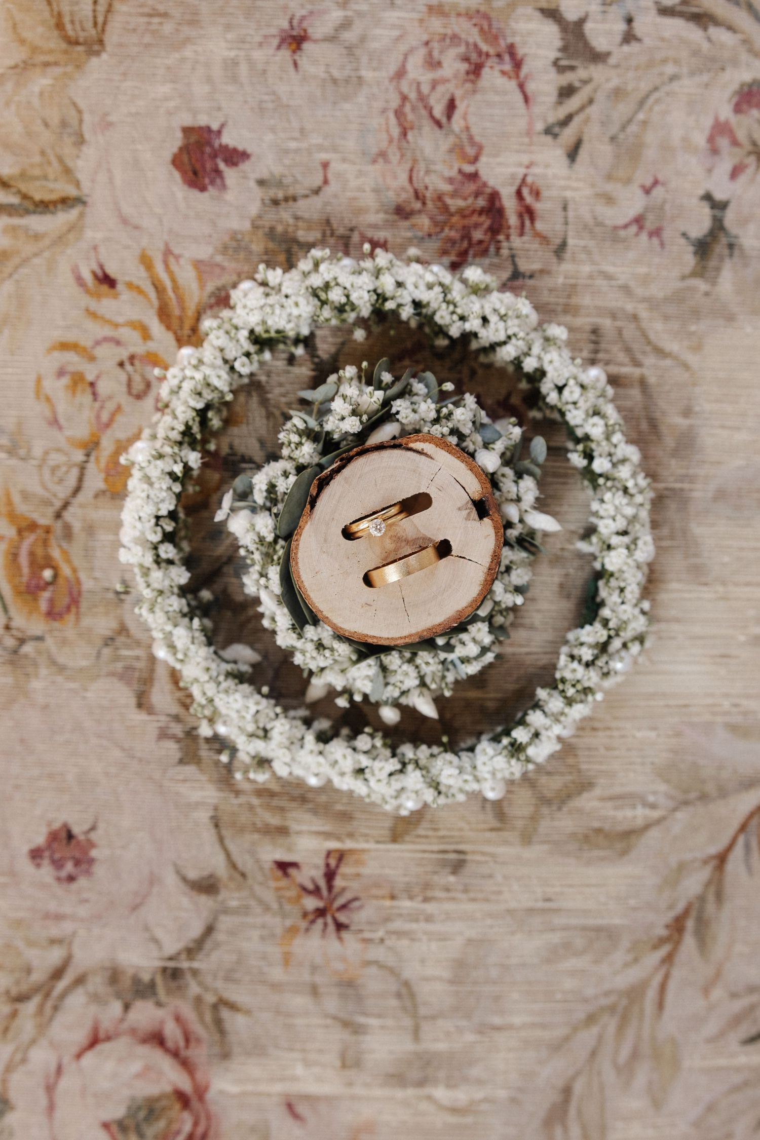 Gold wedding bands on a wood slice surrounded by a baby's breath wreath.