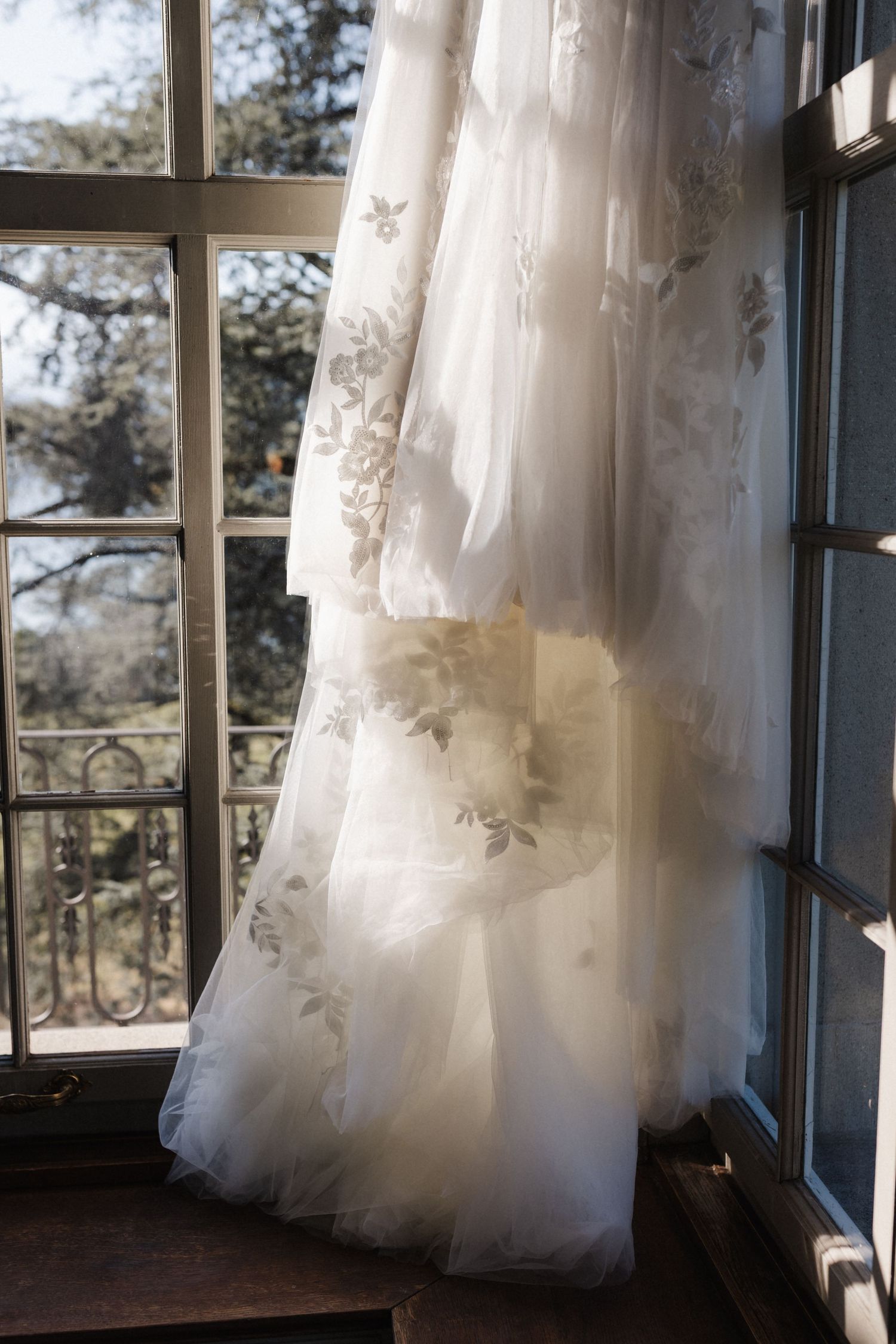Elegant floral tulle wedding gown hanging in sunlit window with outdoor garden backdrop.