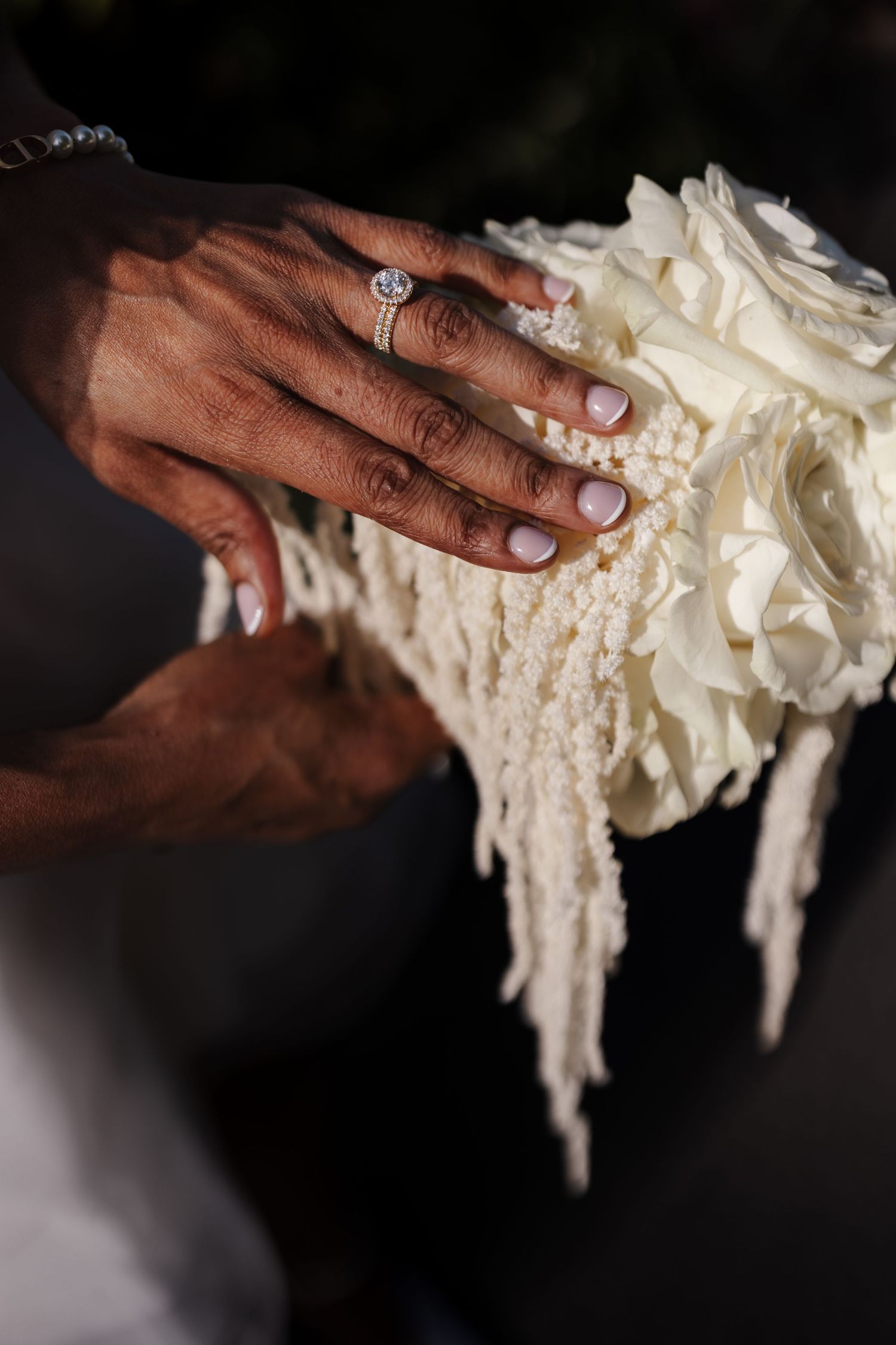 Close-up of a bride's hand with diamond engagement ring resting on a white rose and pearl wedding bouquet.