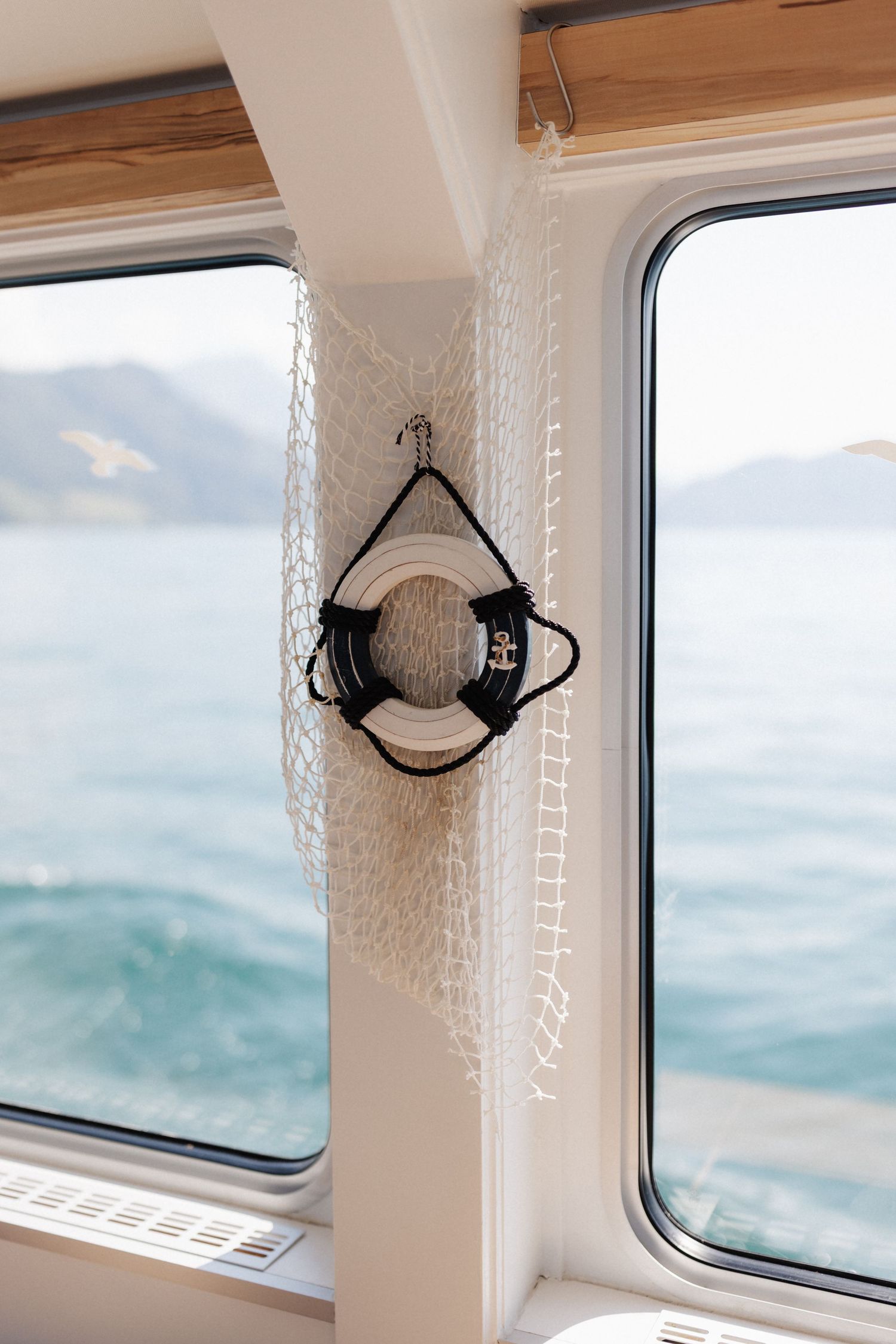 A white life preserver ring hangs on a fishing net between two boat windows overlooking turquoise water and mountains.