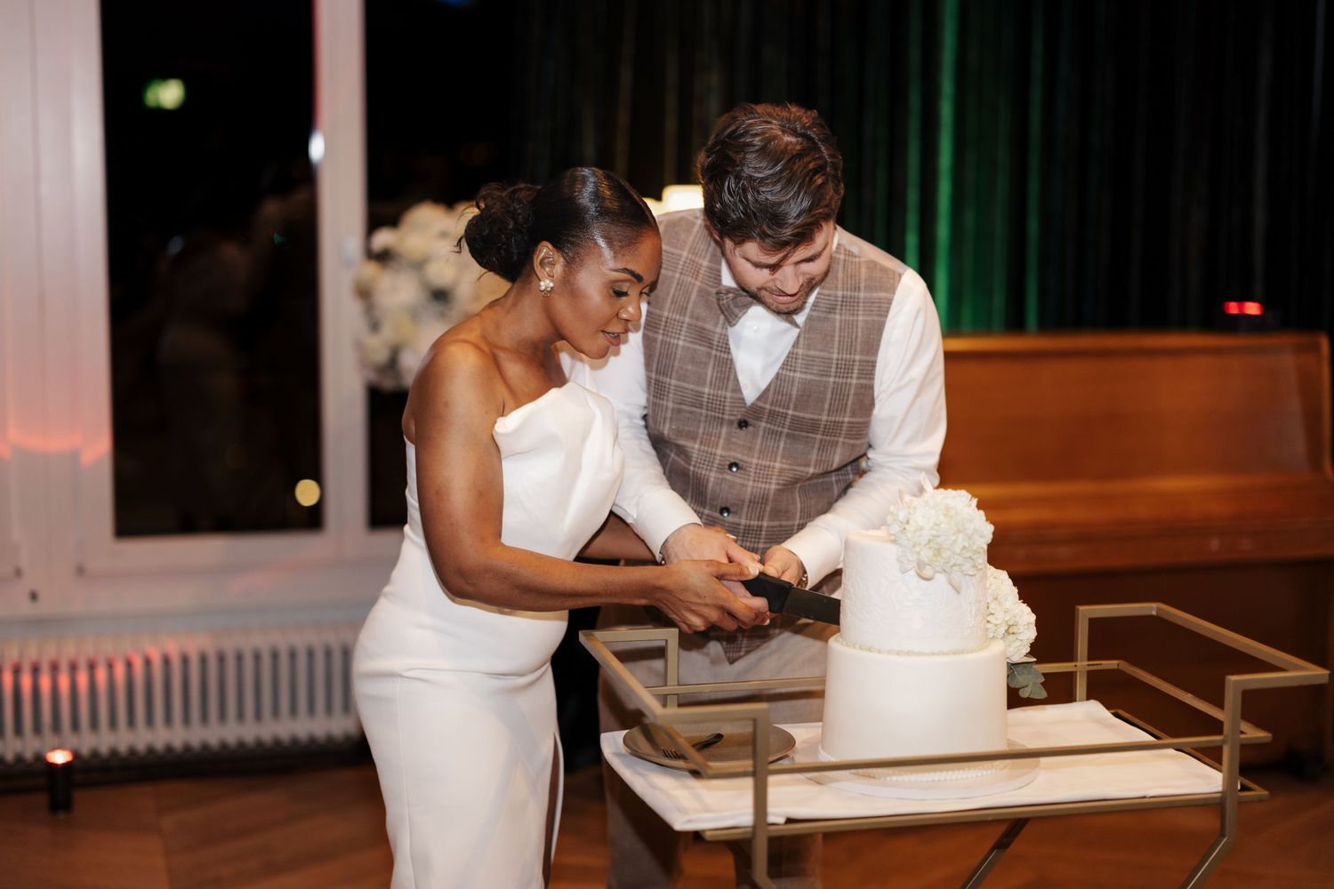 Bride and groom smiling while cutting their white wedding cake at their evening wedding reception.