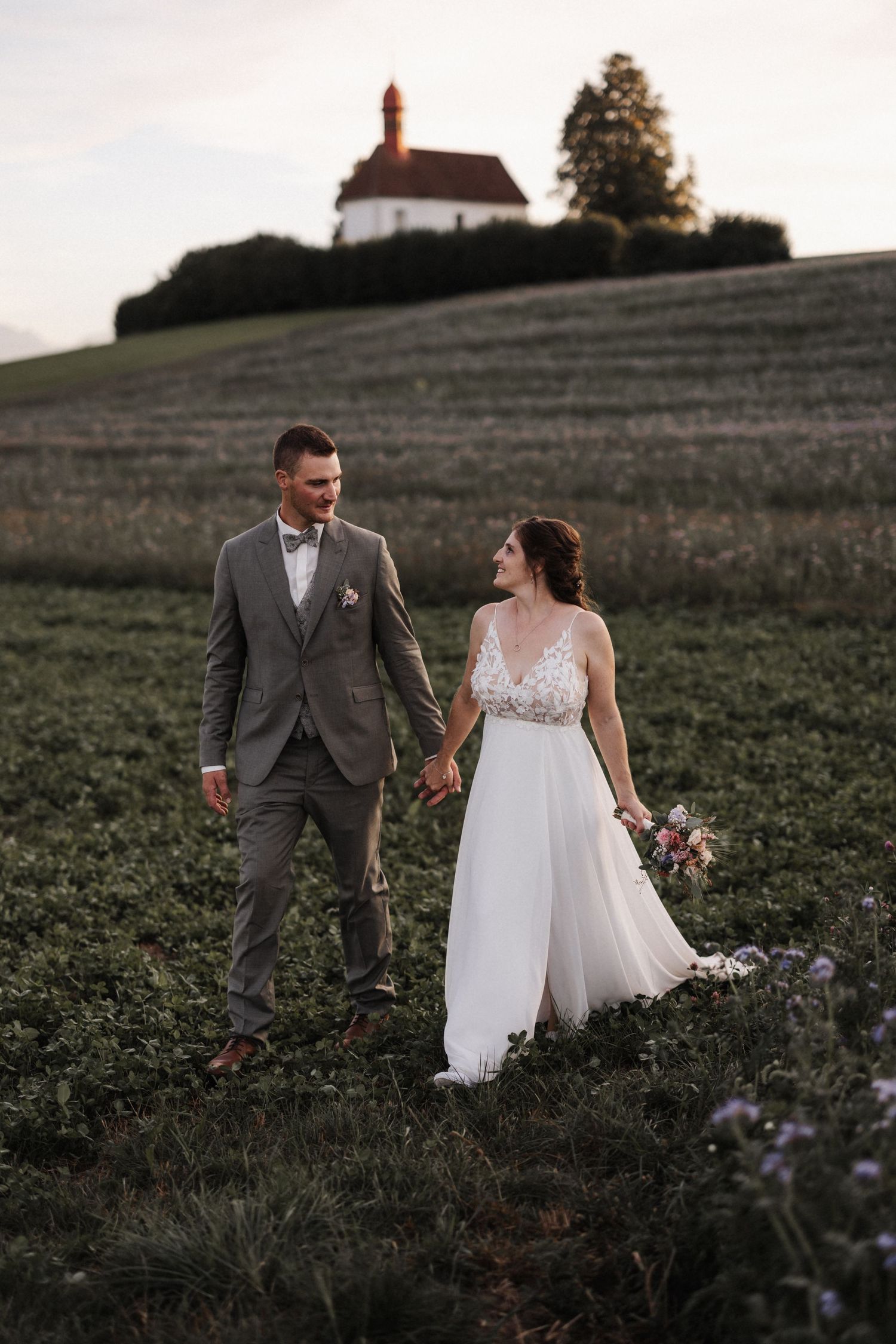 Bride and groom walking hand in hand in a field with a historic chapel on a hill at sunset.