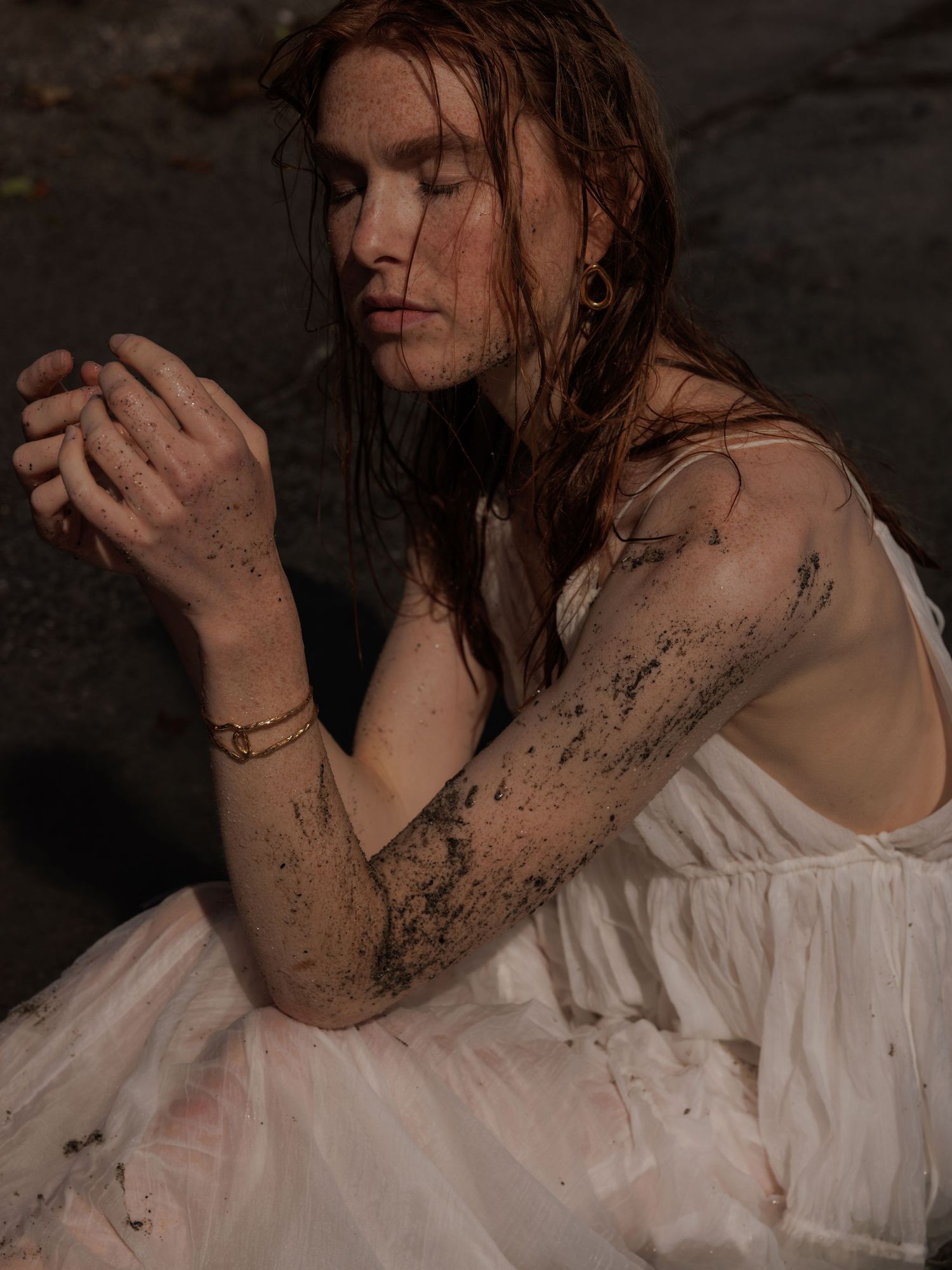 Woman with wet auburn hair covered in mud wearing a white dress, hands clasped in contemplation.