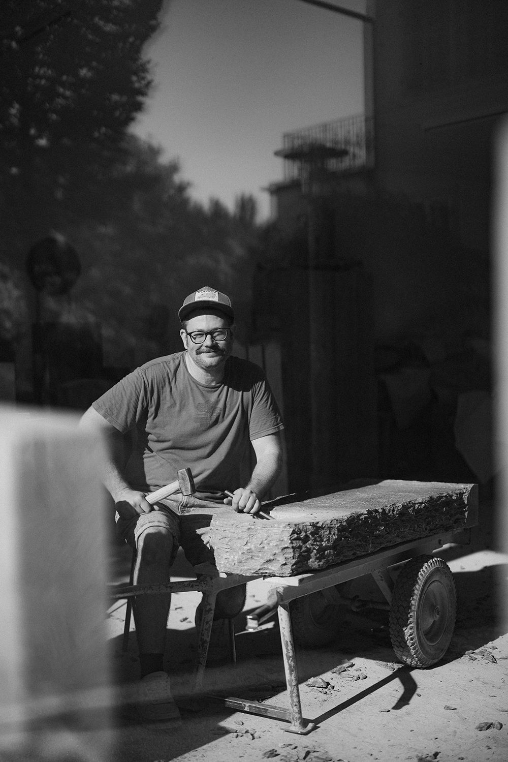 Smiling male sculptor working on a stone slab outdoors in a black and white photograph.