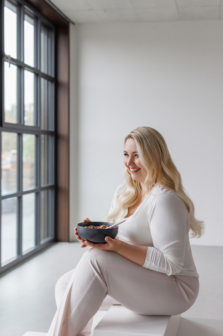 Smiling blonde plus-size woman in white outfit holding dark bowl with berries near large window.