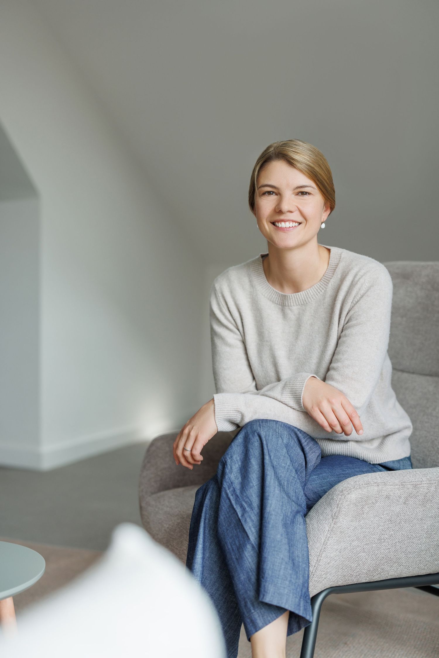 Smiling blonde woman in gray sweater and blue pants sitting in a modern gray armchair.