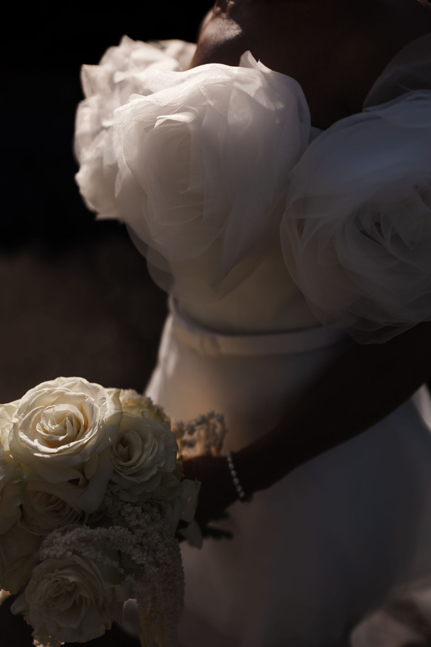 Close-up of bride in white off-shoulder gown with rose appliques holding a white rose bouquet with pearl bracelet.