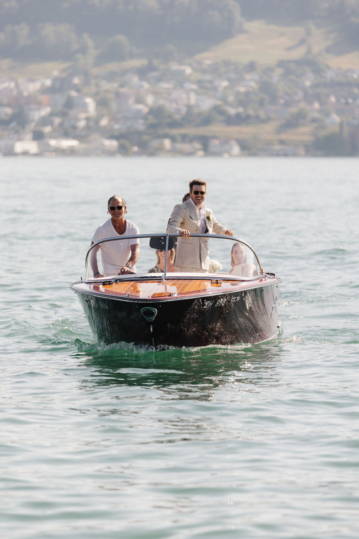 Two men in suits steering a classic wooden speedboat across a scenic alpine lake.