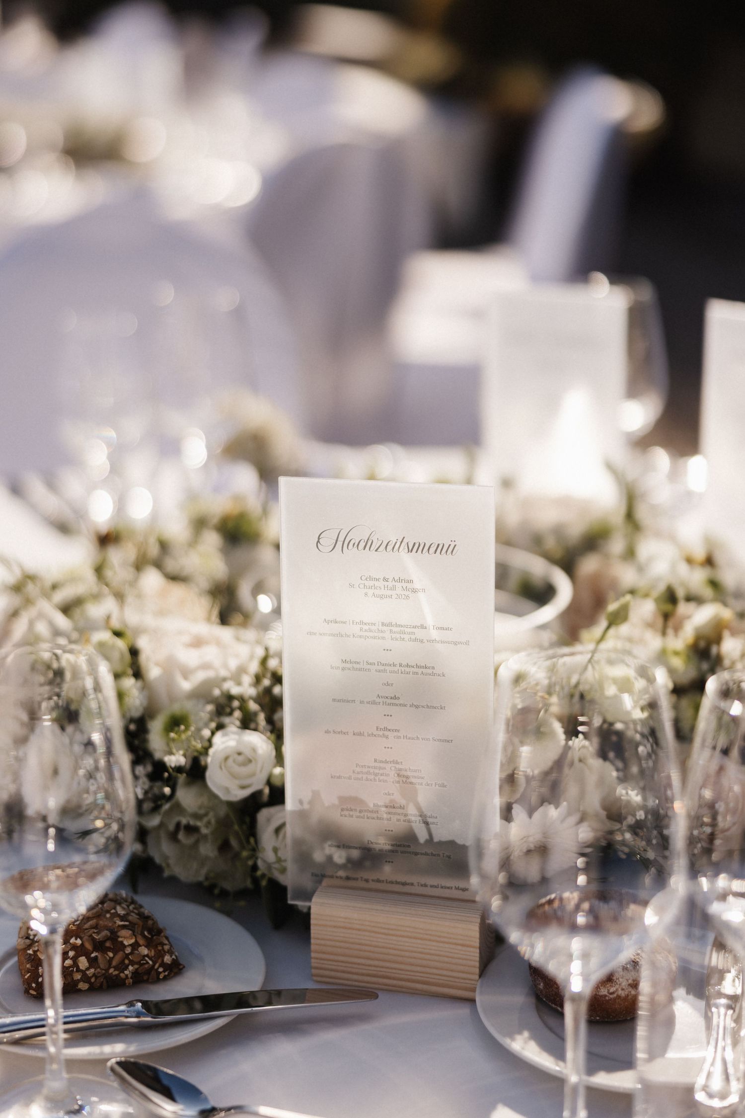 A wedding menu card in a wooden holder sits among white floral arrangements and crystal glasses on a reception table.