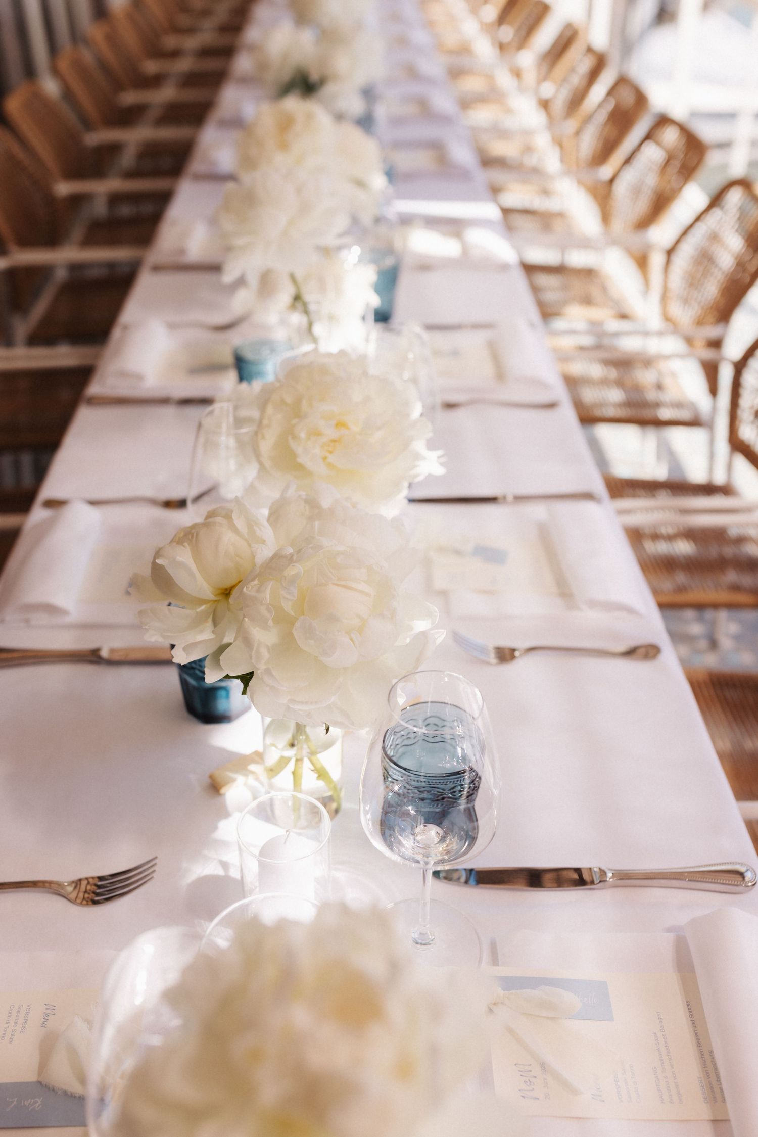 Elegant wedding table runner of white peonies with blue glassware and silver cutlery on crisp white linen.