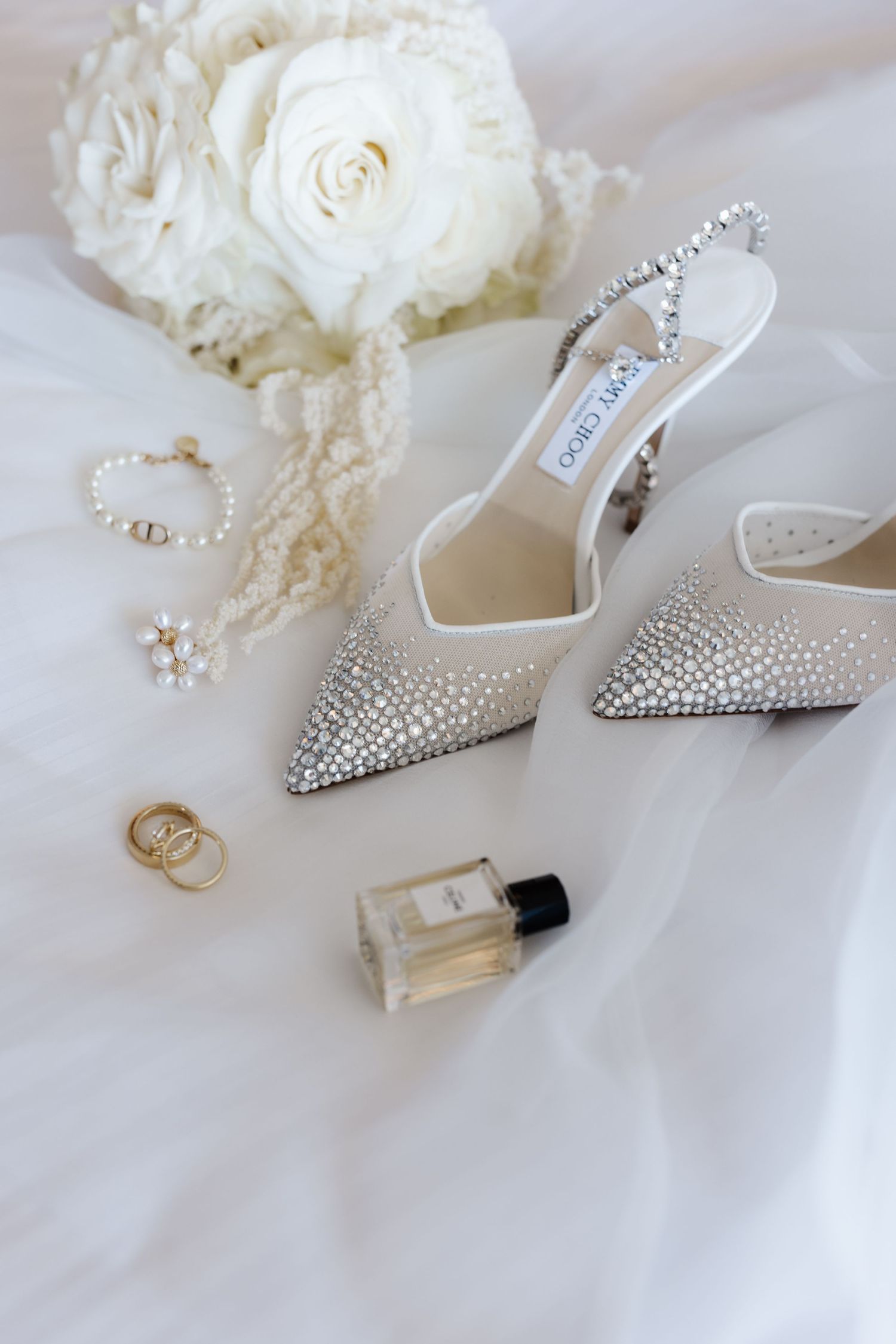 Jimmy Choo crystal slingback bridal heels with gold rings, pearl jewelry, and perfume on white tulle.