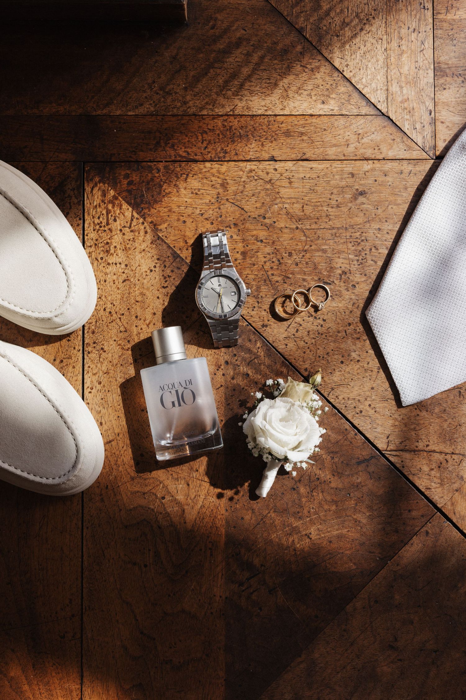 Groom wedding flat lay with Acqua di Gio cologne, silver watch, gold rings, boutonniere and white loafers.