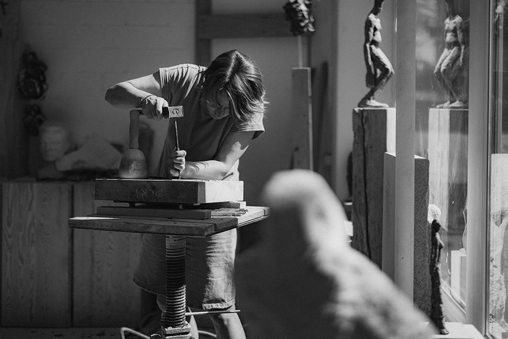 Sculptor works with hammer and chisel on stone in a workshop filled with figurative sculptures.