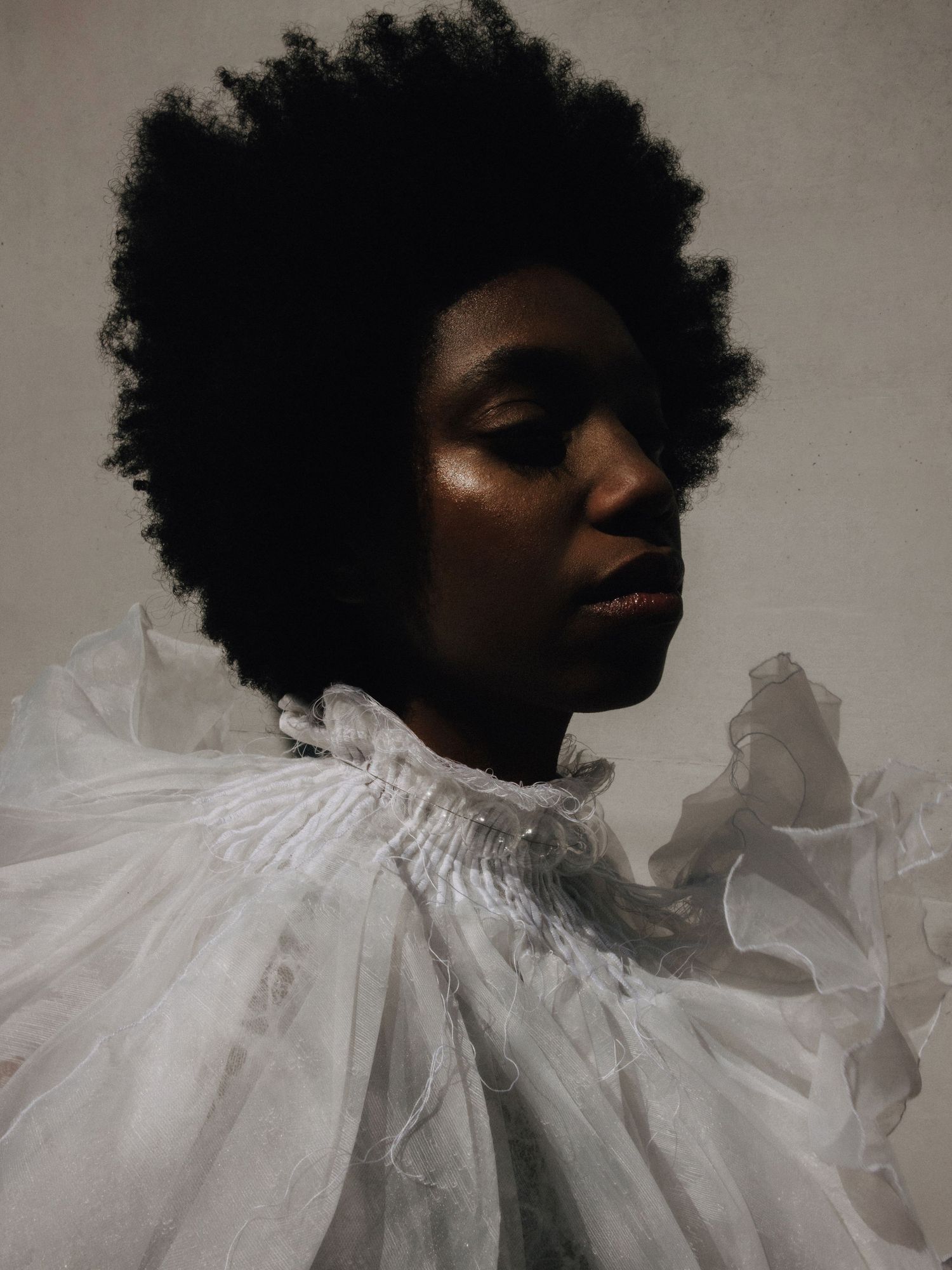 A Black woman with a natural afro wears a dramatic white organza ruffled collar in a moody fashion portrait.
