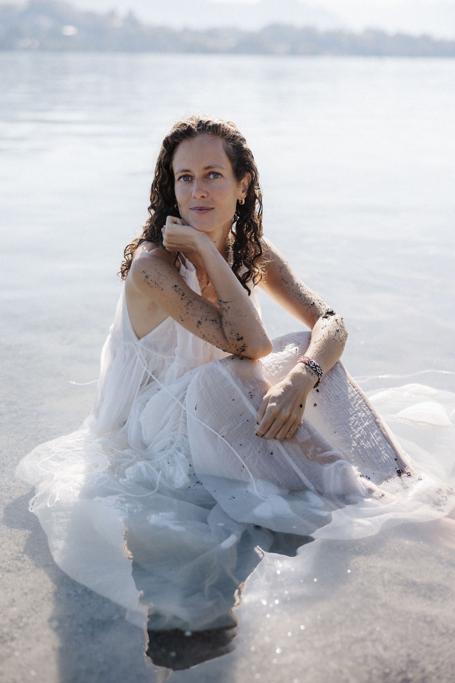 Woman in wet white dress sitting in shallow water, posing with hand on chin, smiling.