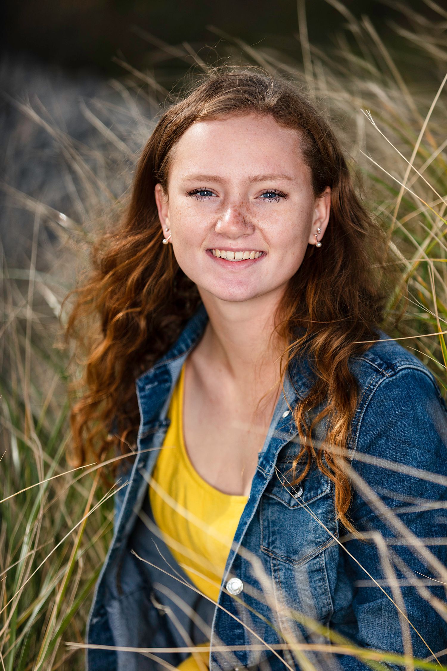 Smiling young woman with red hair wearing a yellow top and denim jacket, sitting among tall grass outdoors.