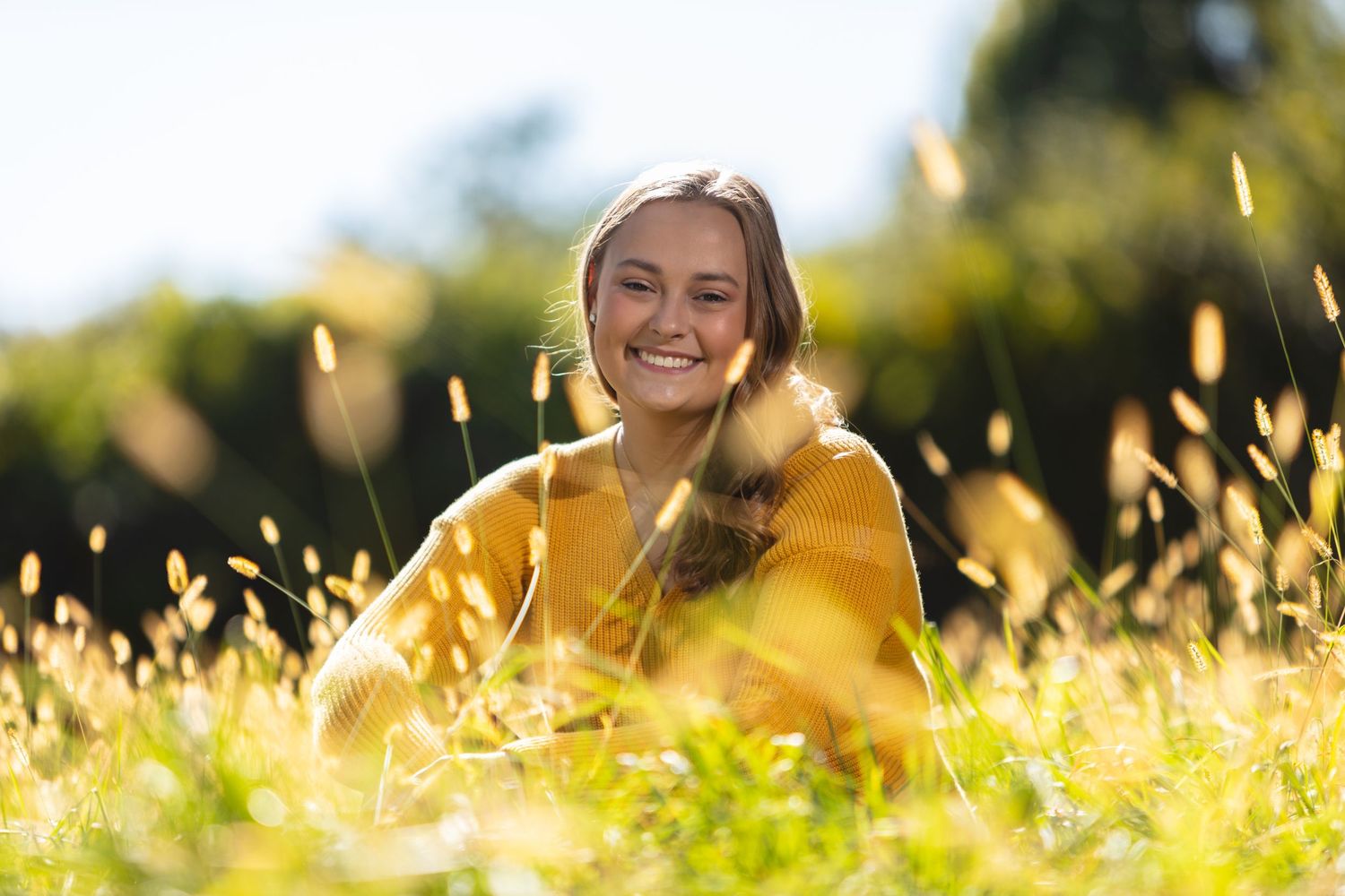 Smiling young woman in yellow sweater sitting in golden sunlit meadow grass outdoors.