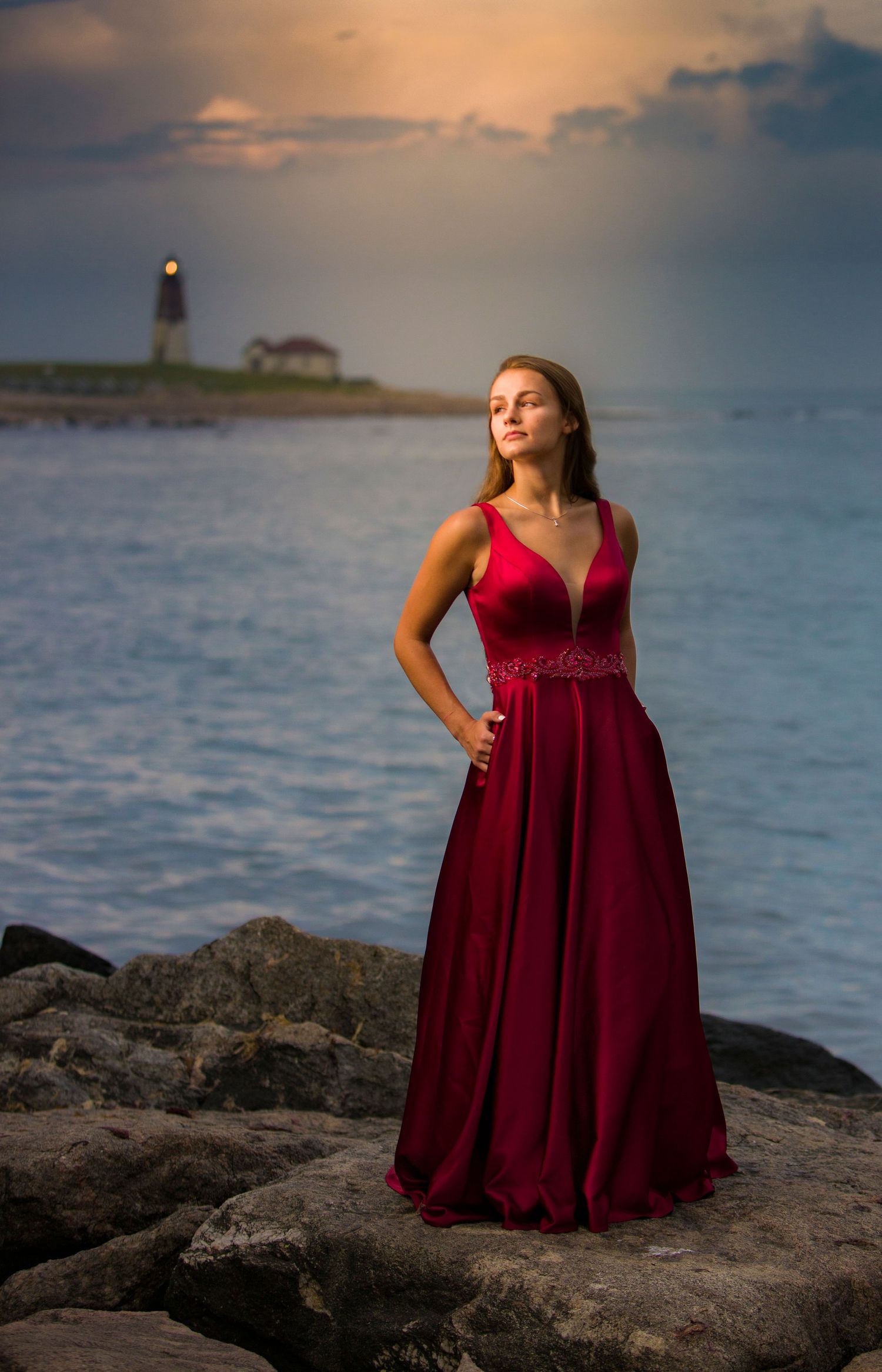 Elegant woman in red gown standing on rocky shore with lighthouse at sunset.