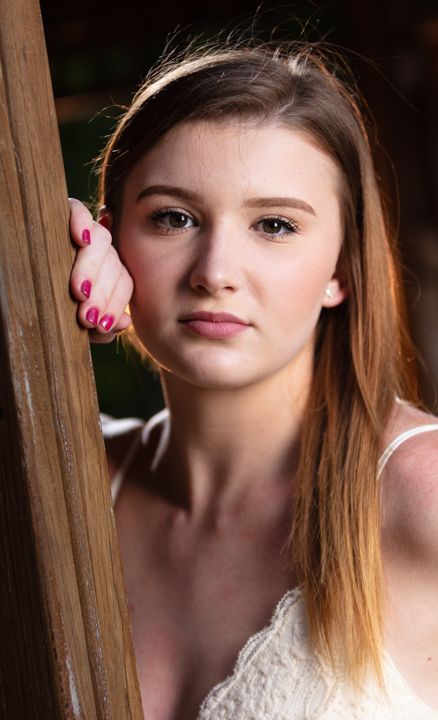 Young woman with long brown hair and pink nails leans against a wooden post in a portrait photo.