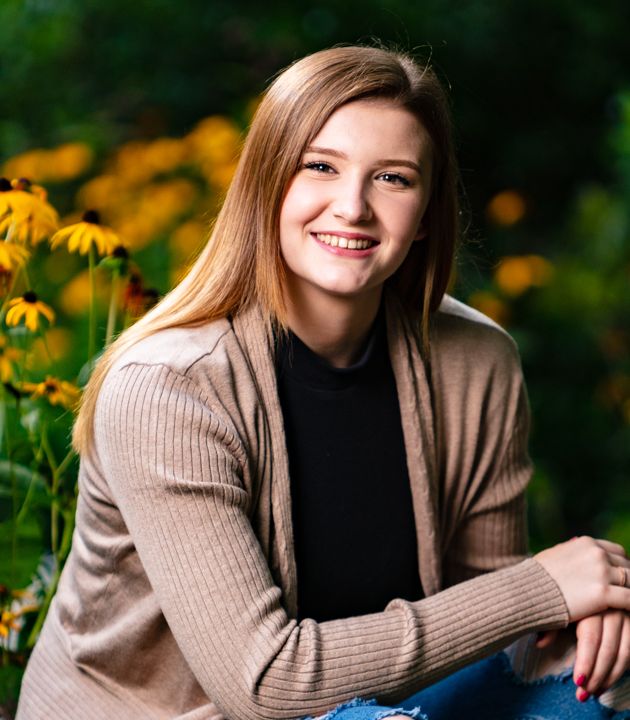 Smiling teenage girl with long blonde hair wearing a beige cardigan, seated outdoors among yellow flowers.
