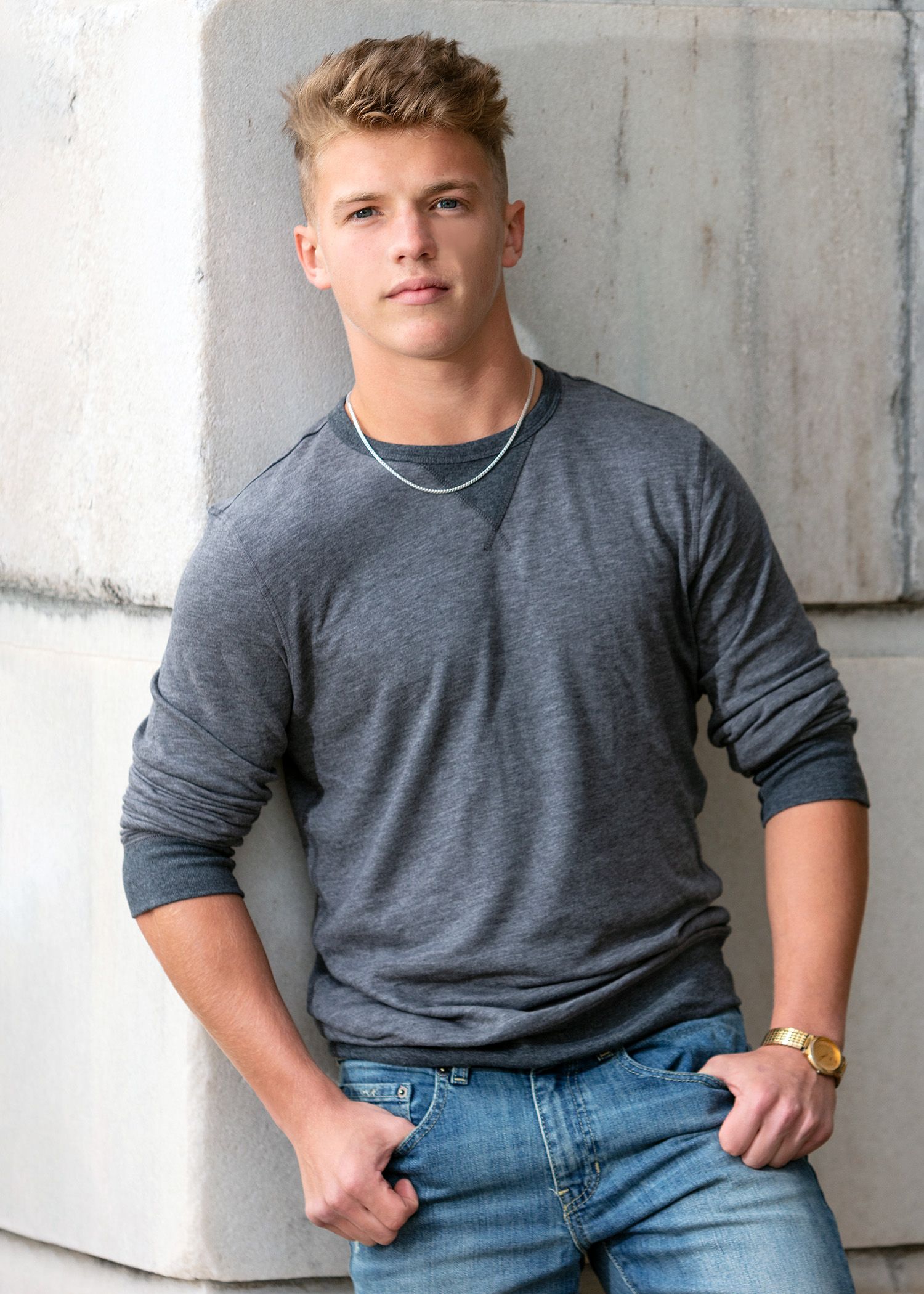 Young athletic man in gray long-sleeve shirt and jeans leaning against concrete wall wearing gold watch.