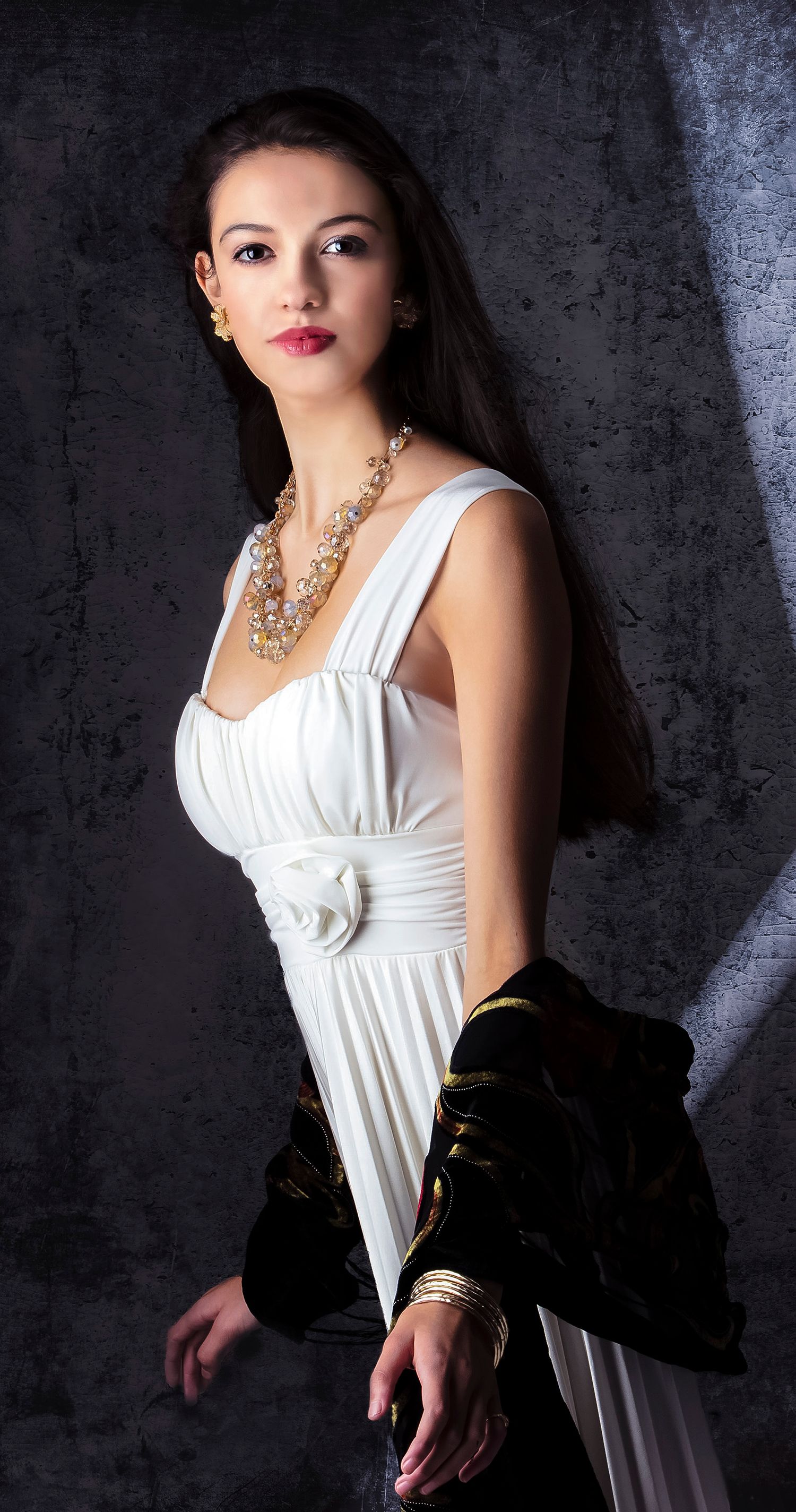 Young woman posing in elegant white dress with gold jewelry against dark studio background.