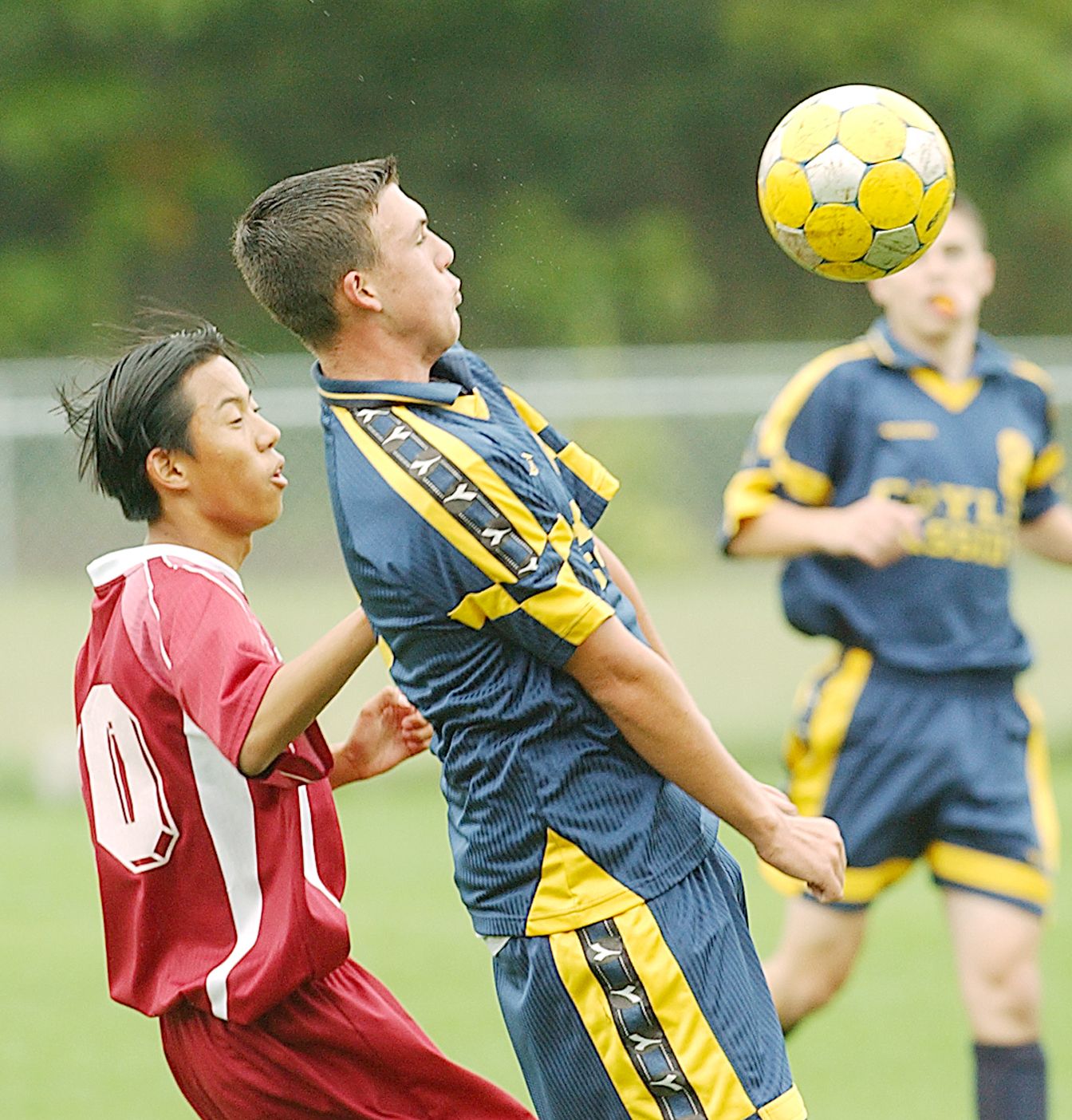 Two youth soccer players compete for a header during a match, with a yellow and black ball in the air.
