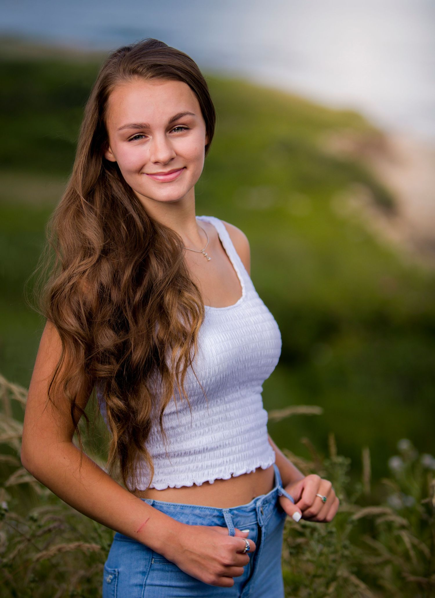 Young woman with long wavy brown hair in white crop top and jeans posing in green field.