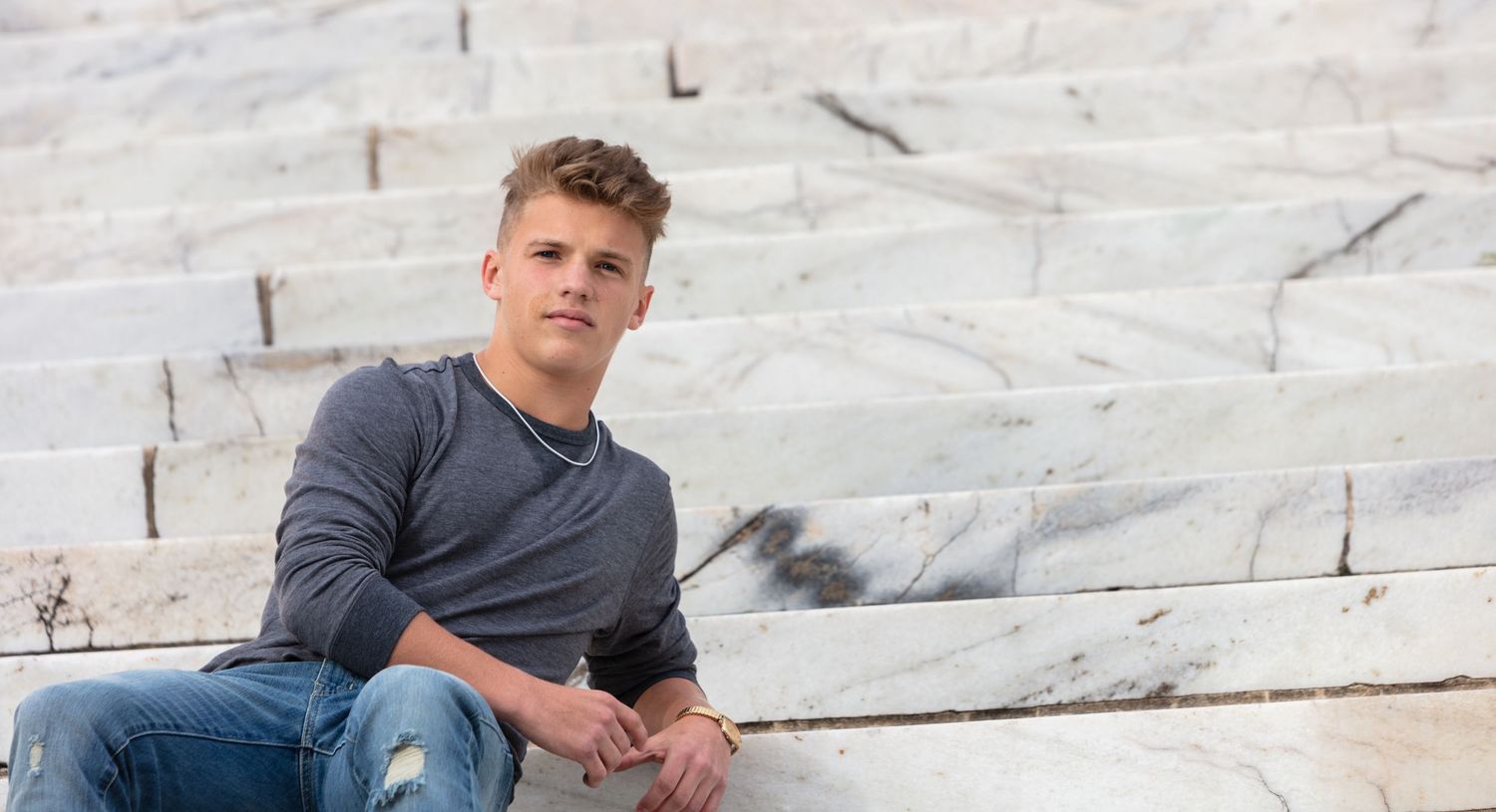 Young man in gray sweater sitting on marble steps in casual outdoor senior portrait photo.