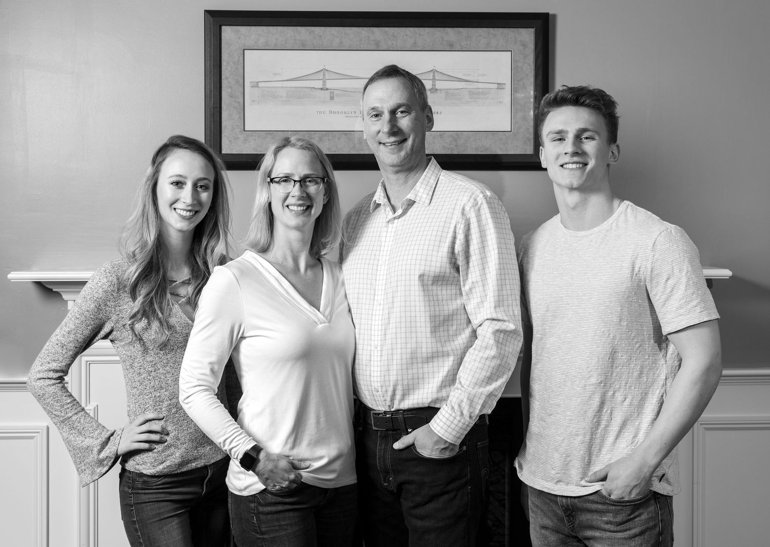 Black and white portrait of happy family of four posing indoors by fireplace mantel.