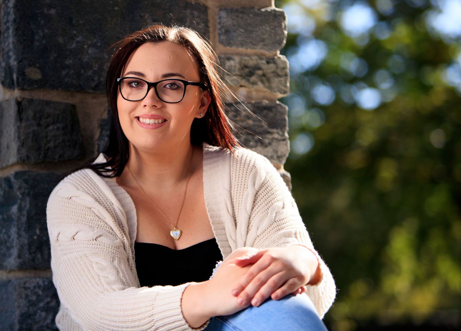 Smiling young woman in cardigan sits against stone wall outdoors in natural light.