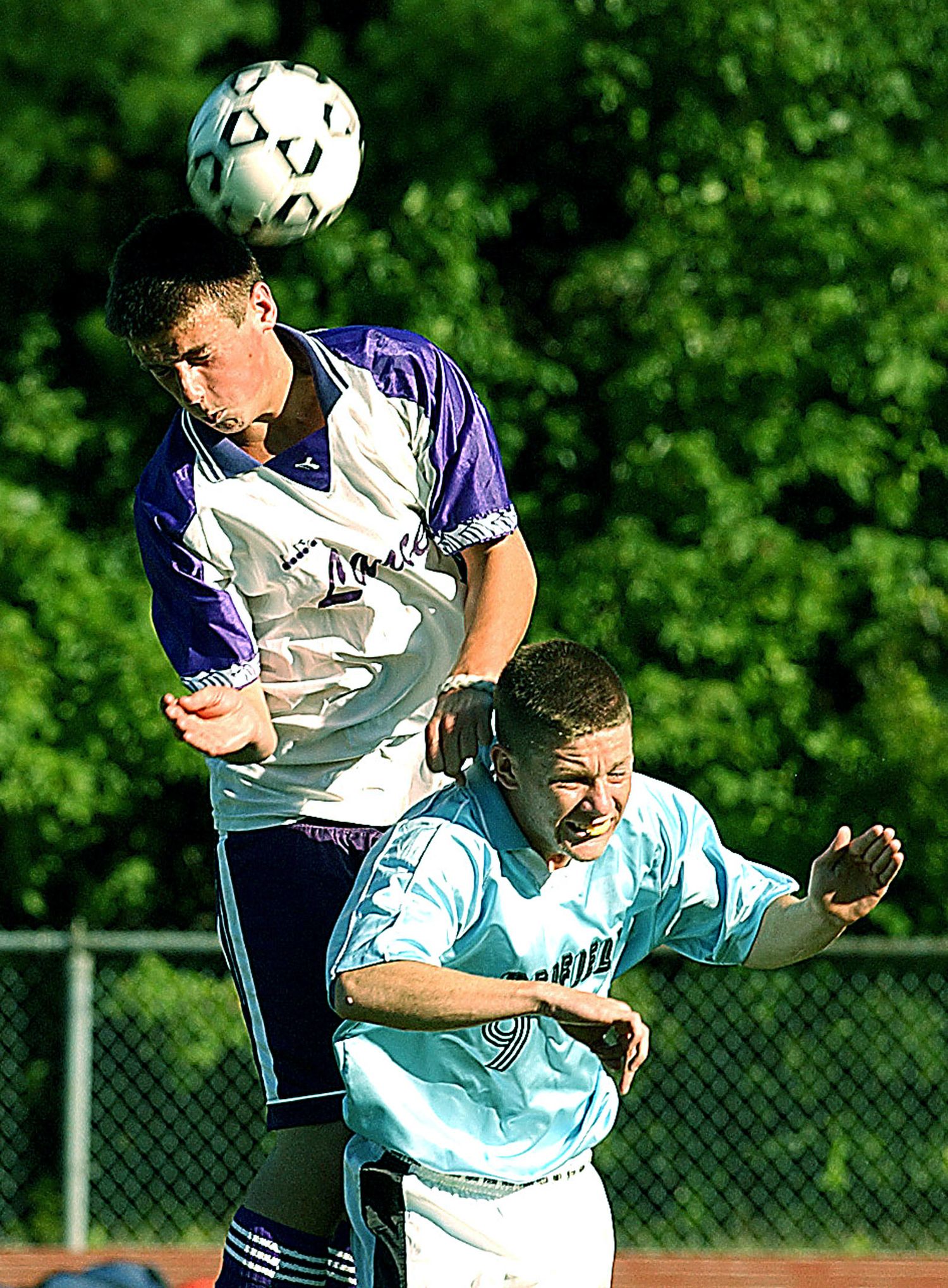 Two soccer players compete for a header ball during an outdoor match, one player in purple leaping above the other in light blue.