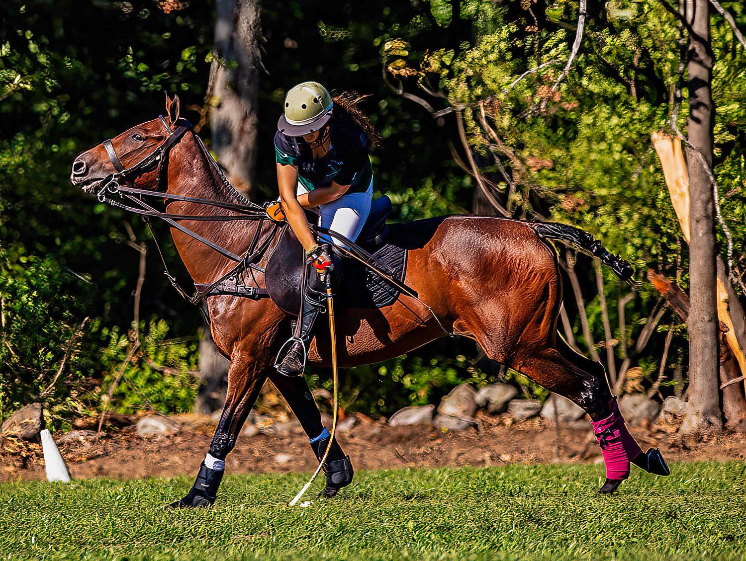 Polo player in green helmet rides chestnut horse, leaning down with mallet during competitive match on lush green field.